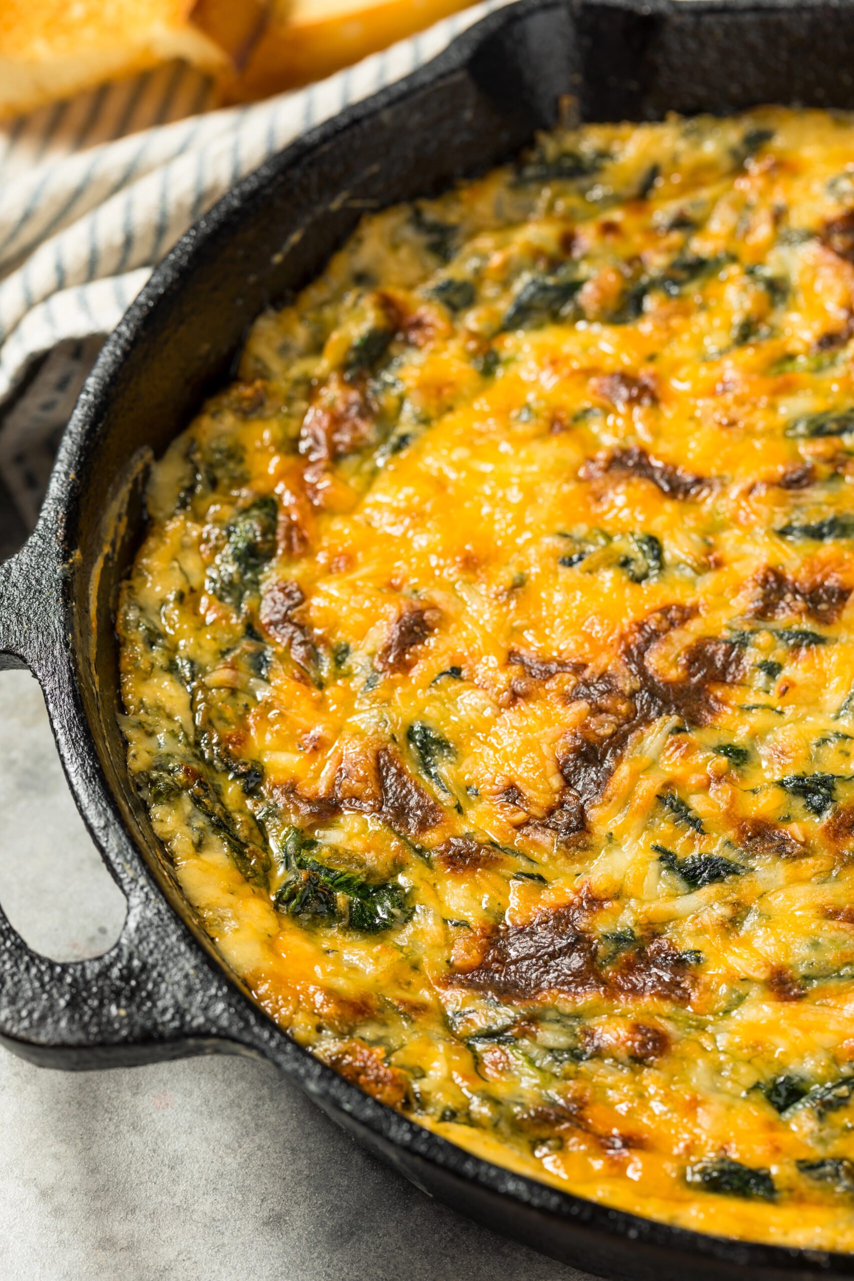 Creamy baked Spinach Maria Dip in a cast iron skillet topped with melted cheddar and mozzarella cheese, ready to serve with bread and chips.