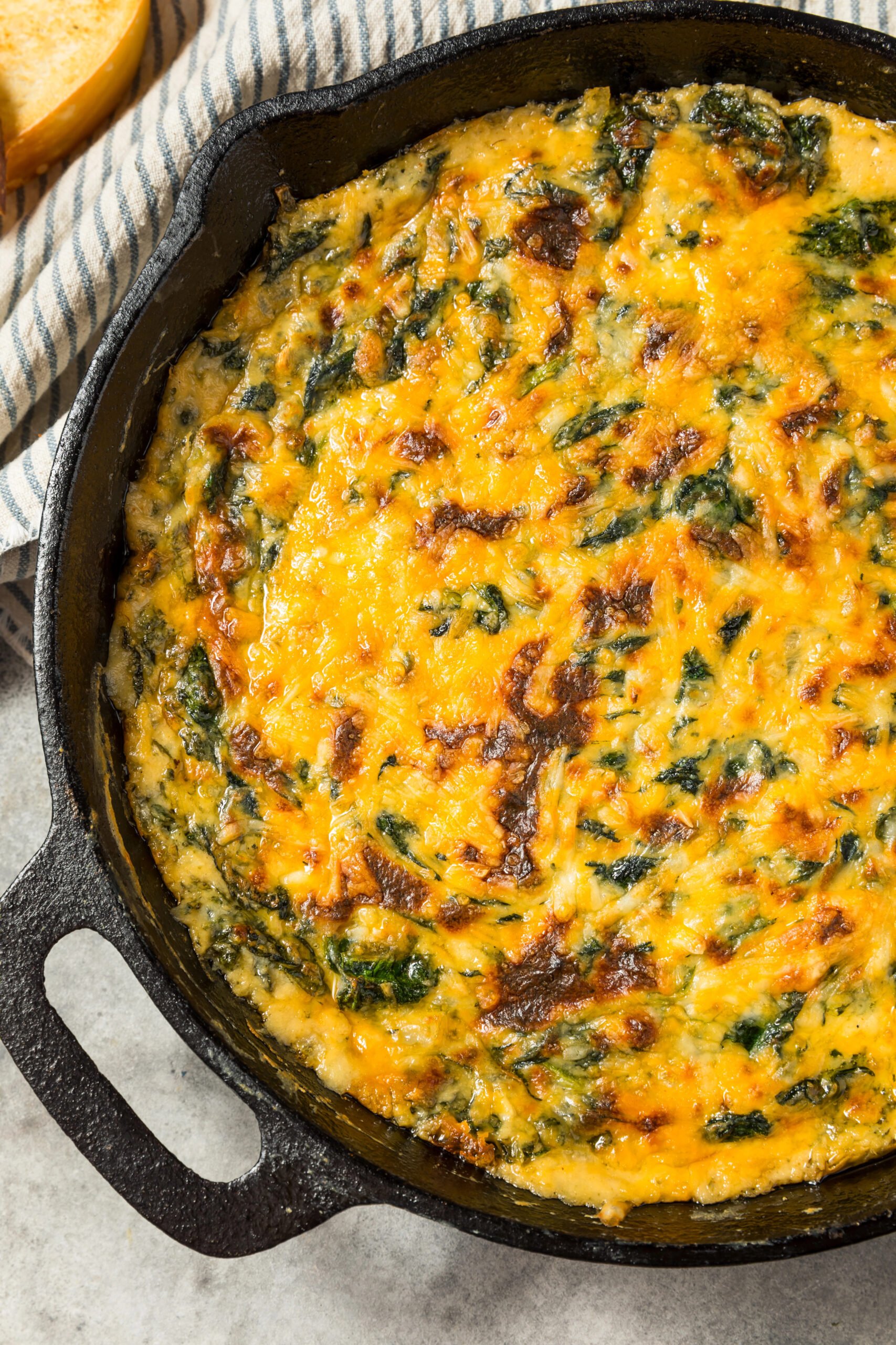 Creamy baked Spinach Maria Dip in a cast iron skillet topped with melted cheddar and mozzarella cheese, ready to serve with bread and chips.