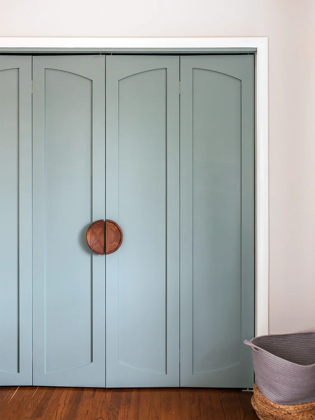 Soft blue bifold closet doors with arched panel detailing and a large round wooden handle, set against white walls and hardwood floors.