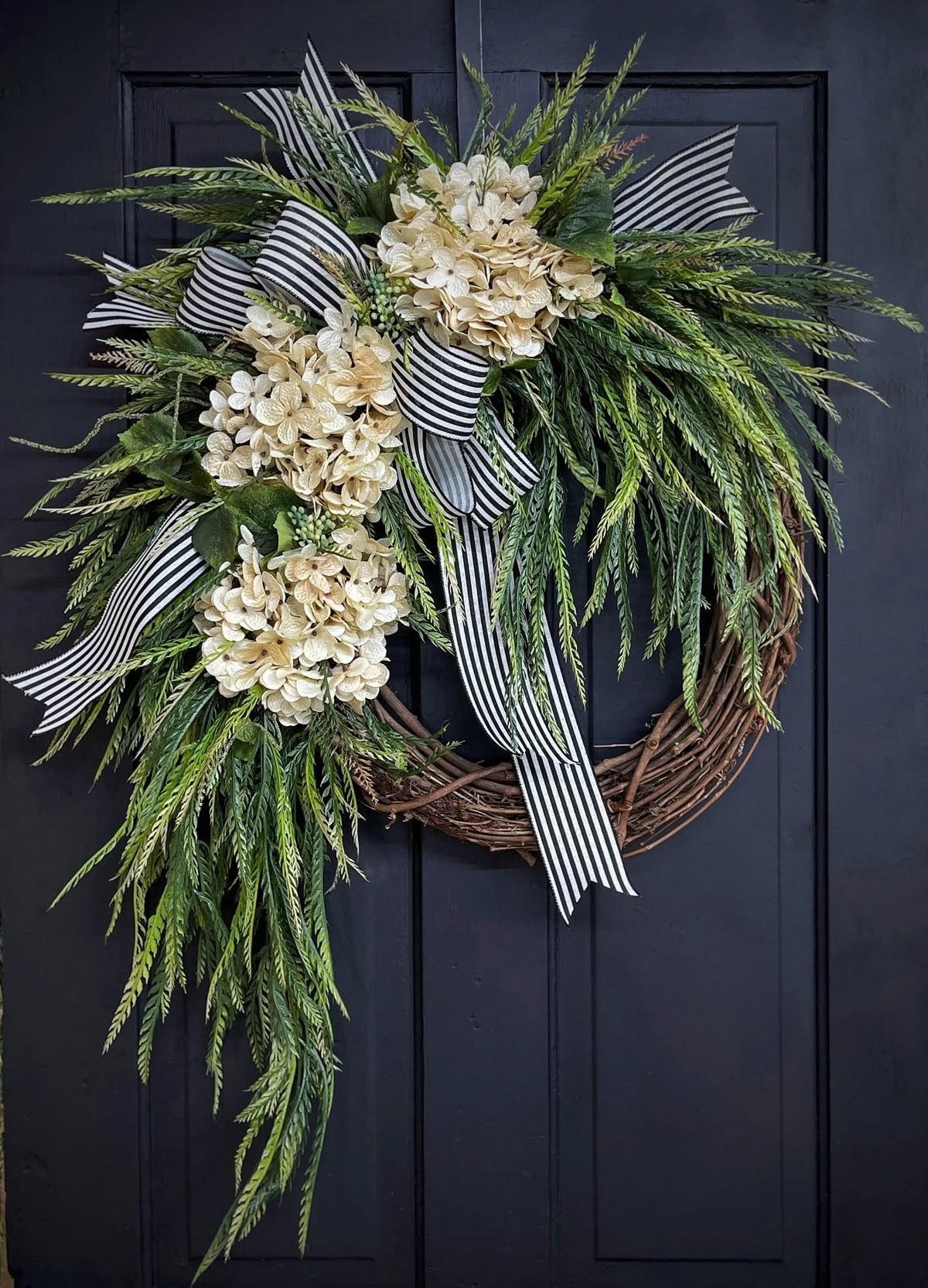 Asymmetrical front door wreath with cascading greenery, cream hydrangeas, and a black and white striped ribbon on a dark door