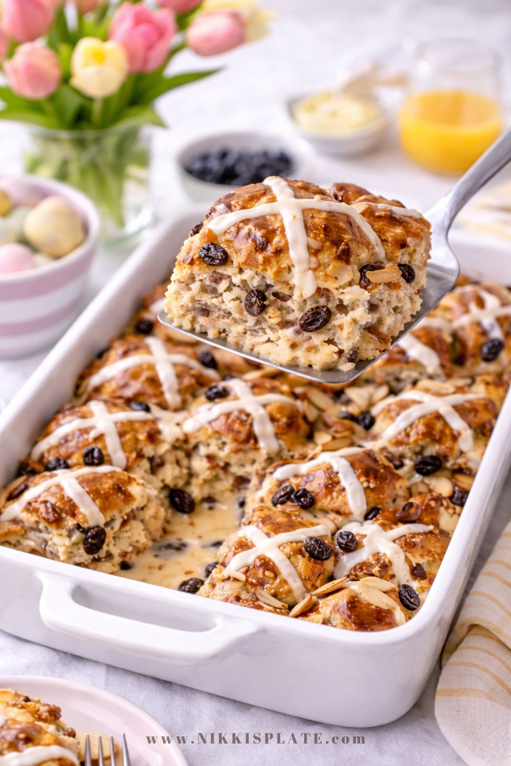 A slice of hot cross bun French toast casserole being lifted from a white baking dish, showing the creamy custard interior with raisins, toasted almonds, and icing drizzle, styled on an Easter brunch table with tulips and pastel decor.