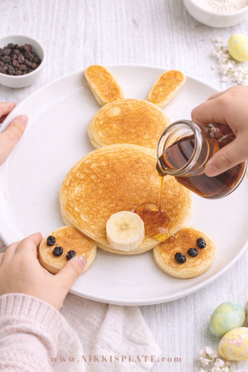 Child's hands decorating sourdough Easter bunny pancakes with banana slices and raisins on a white plate