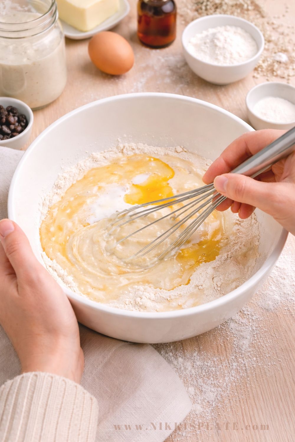 Woman's hands whisking sourdough pancake batter in a white bowl with flour, egg, butter, and vanilla on a wooden countertop for Easter bunny pancakes recipe