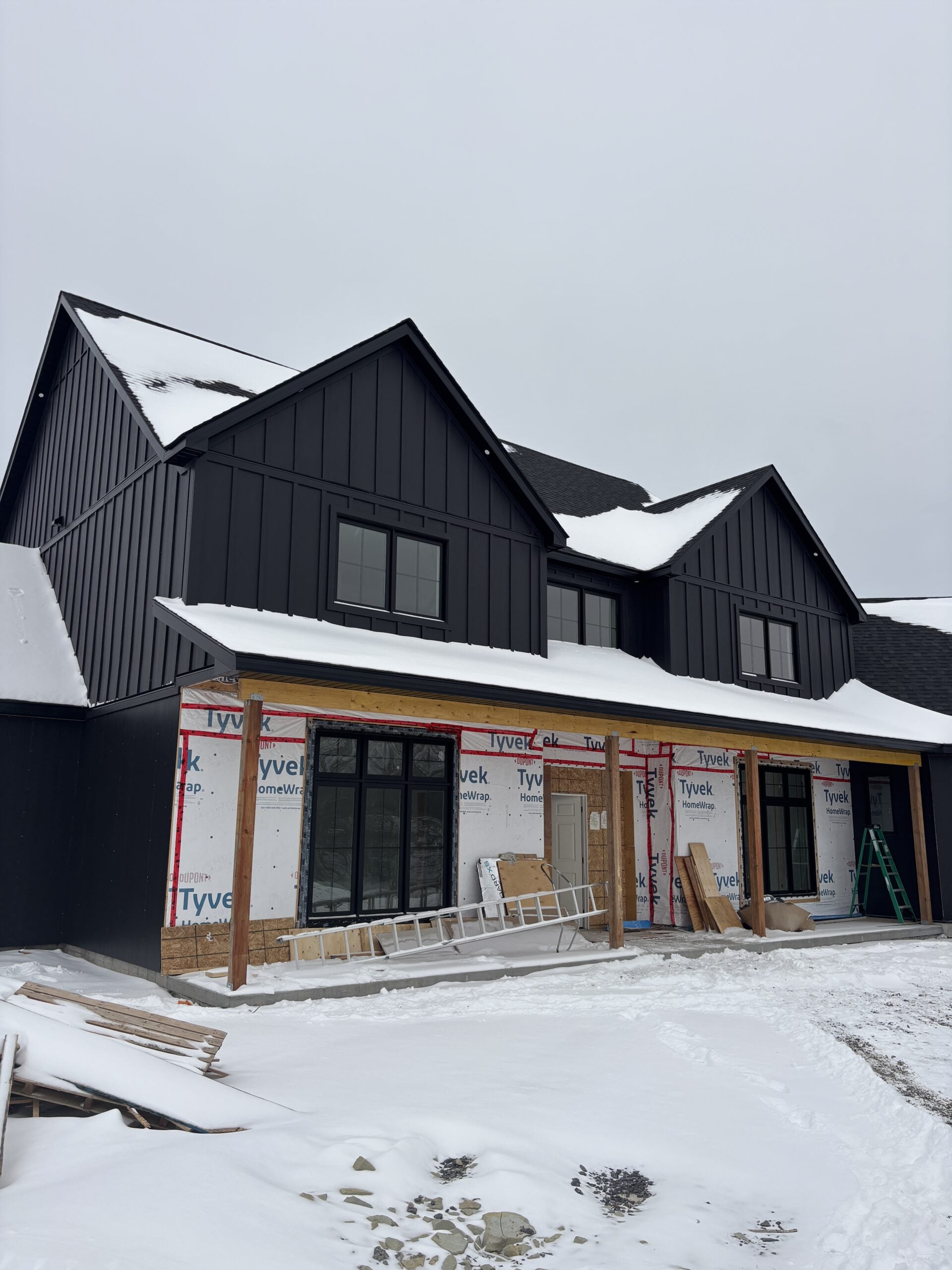 Modern farmhouse exterior with black board and batten siding, black windows, and covered front porch during winter construction on our rural Ontario build.