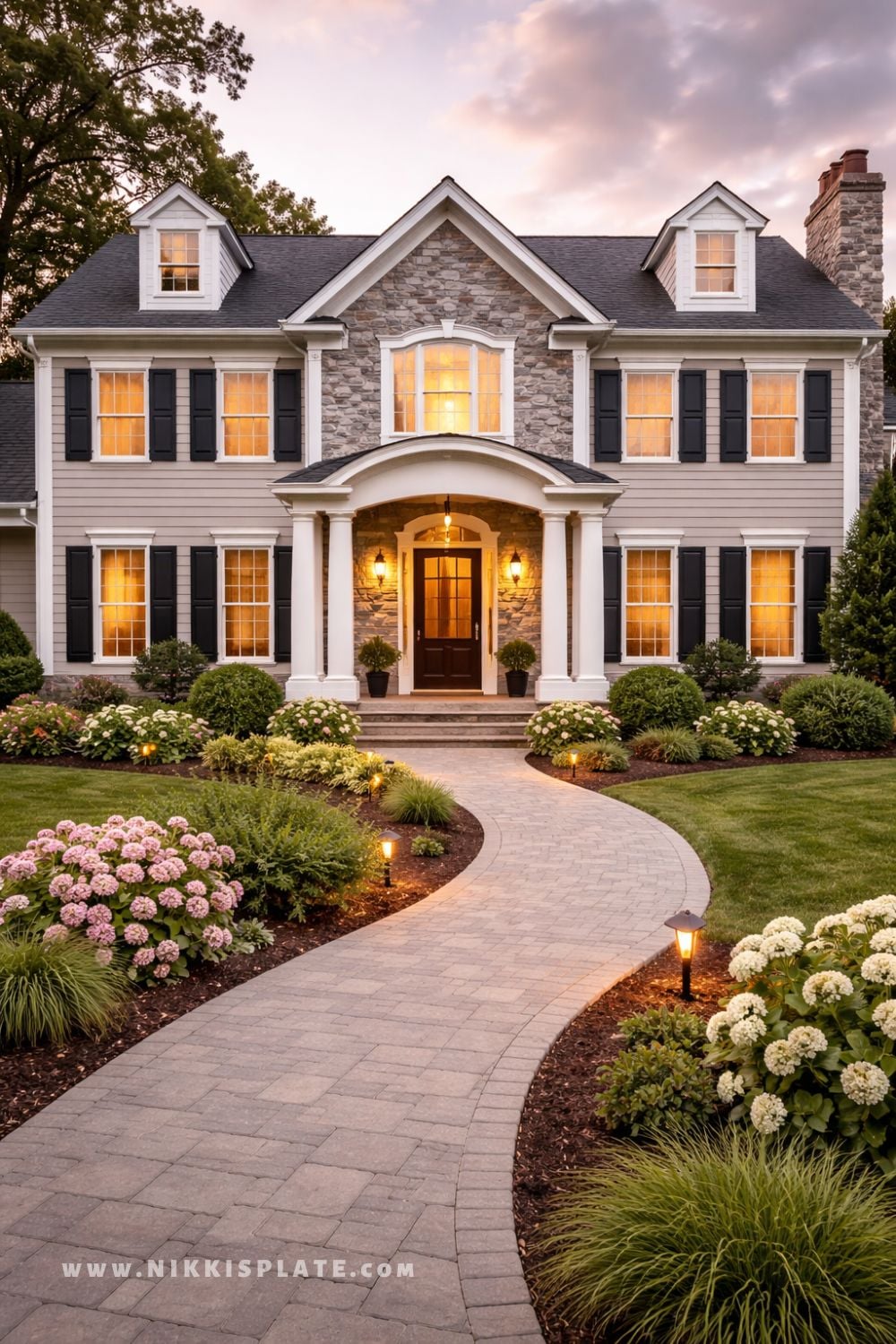 Traditional two-story home exterior with stone accents, black shutters, white columns, and landscaped front walkway at sunset