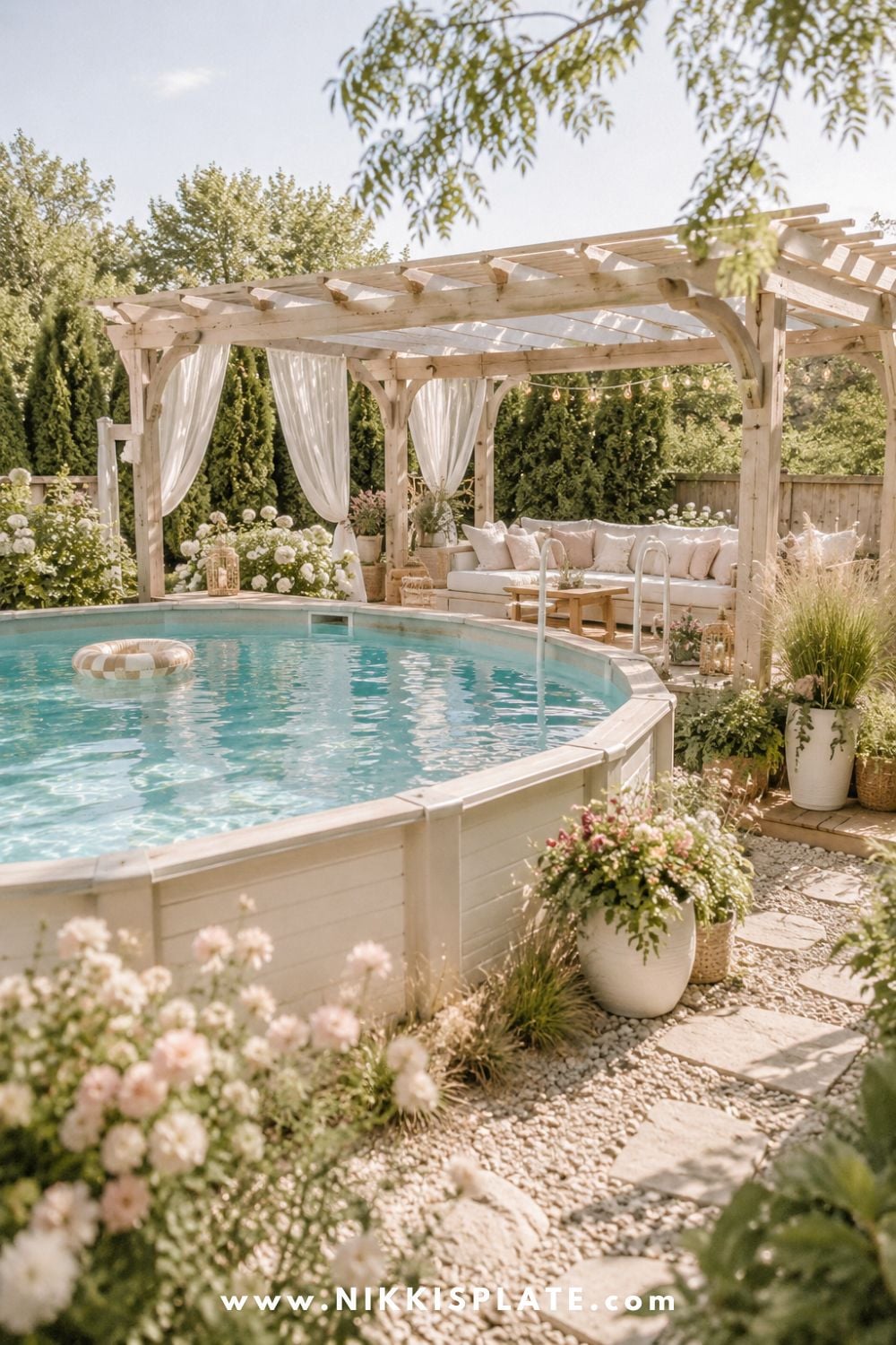 Above ground pool with a light wood pergola, soft white curtains, cozy seating, potted flowers, and a bright gravel walkway.