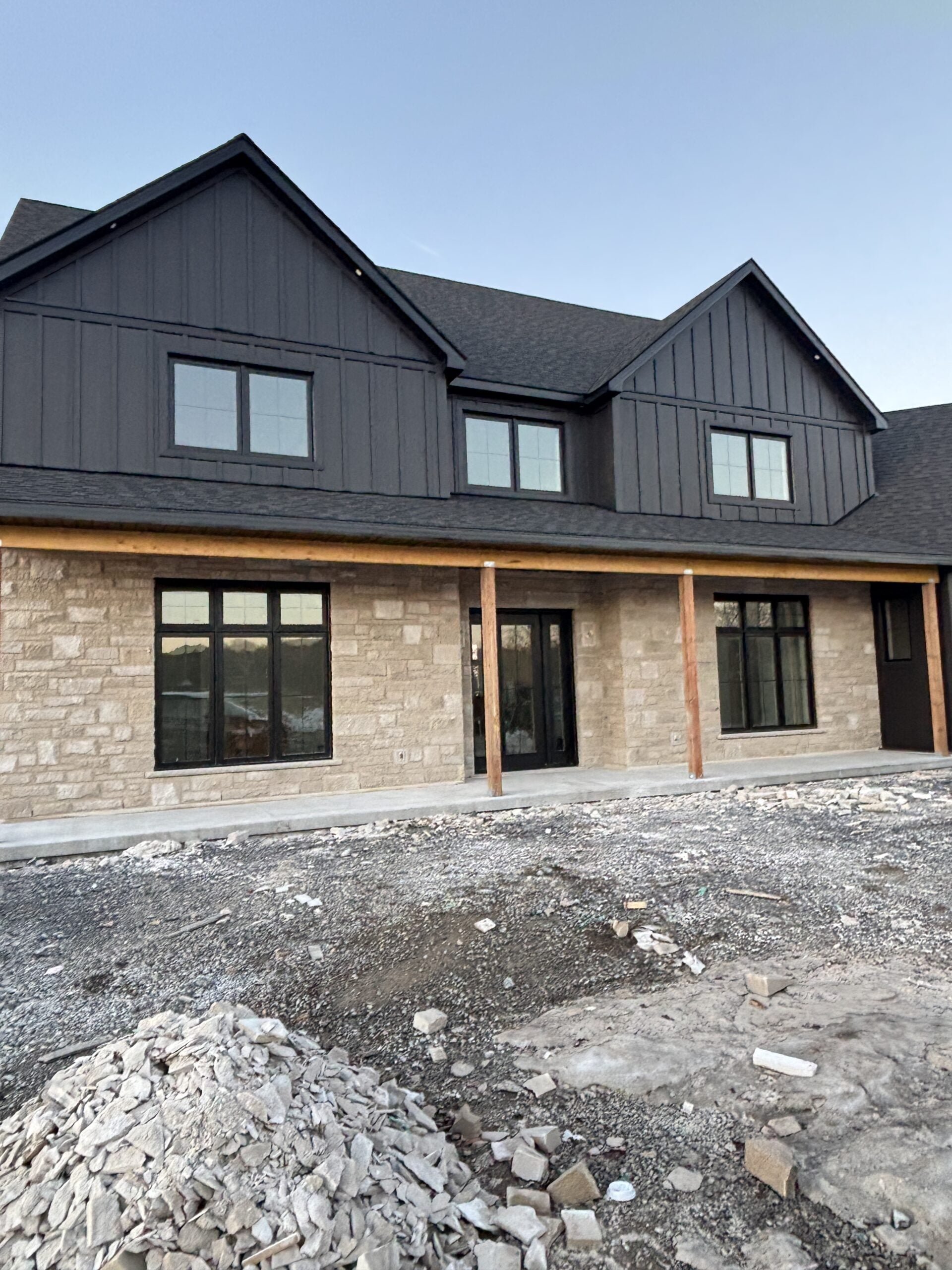 Modern farmhouse exterior featuring limestone front, black board and batten siding, black windows, and covered porch during construction on our rural Ontario custom home build.