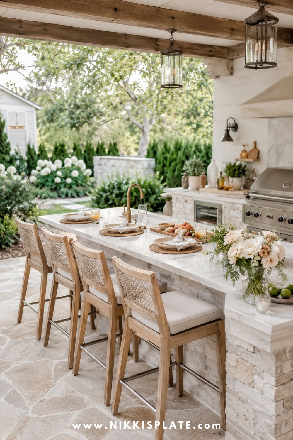 Outdoor kitchen and bar ideas for a backyard with stone island, wood bar seating, built-in grill, lantern lights, flowers, and cozy farmhouse patio decor.