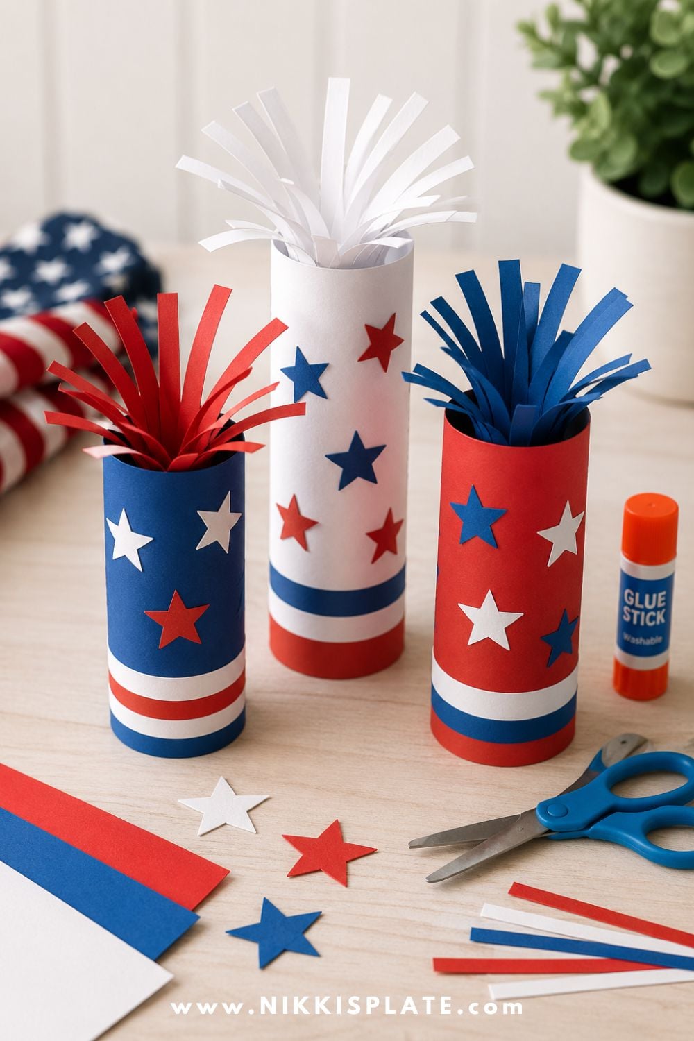 Paper firecracker craft for kids with red, white, and blue paper tubes, paper sparks, star cutouts, glue stick, and scissors on a light craft table