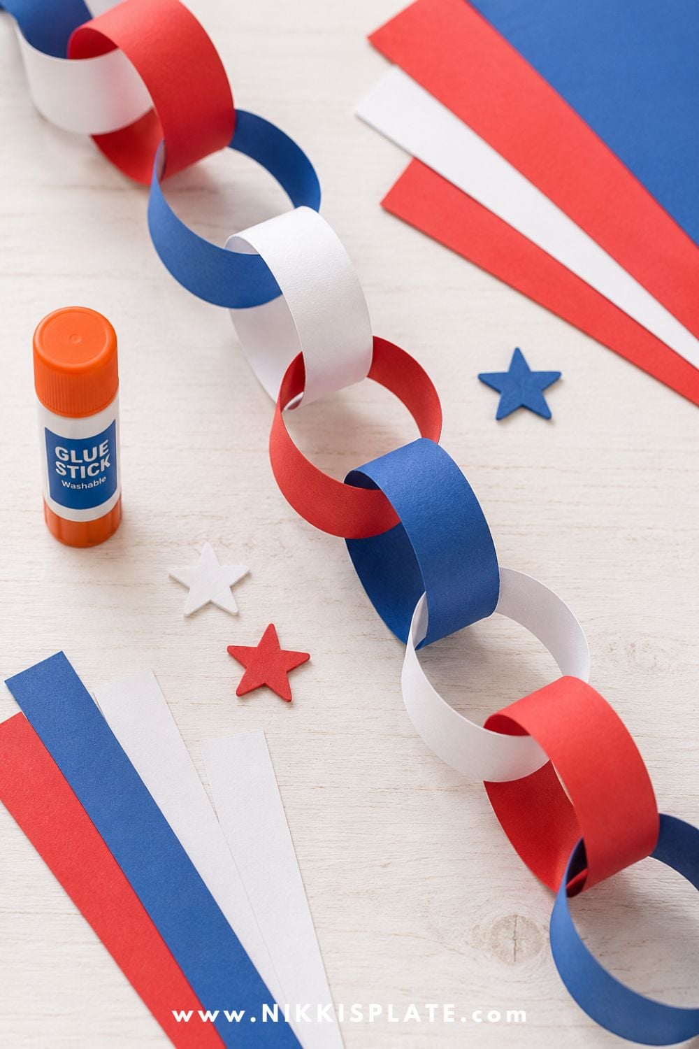 Red, white and blue paper chain craft for kids with construction paper strips, glue stick, and star decorations on a light craft table