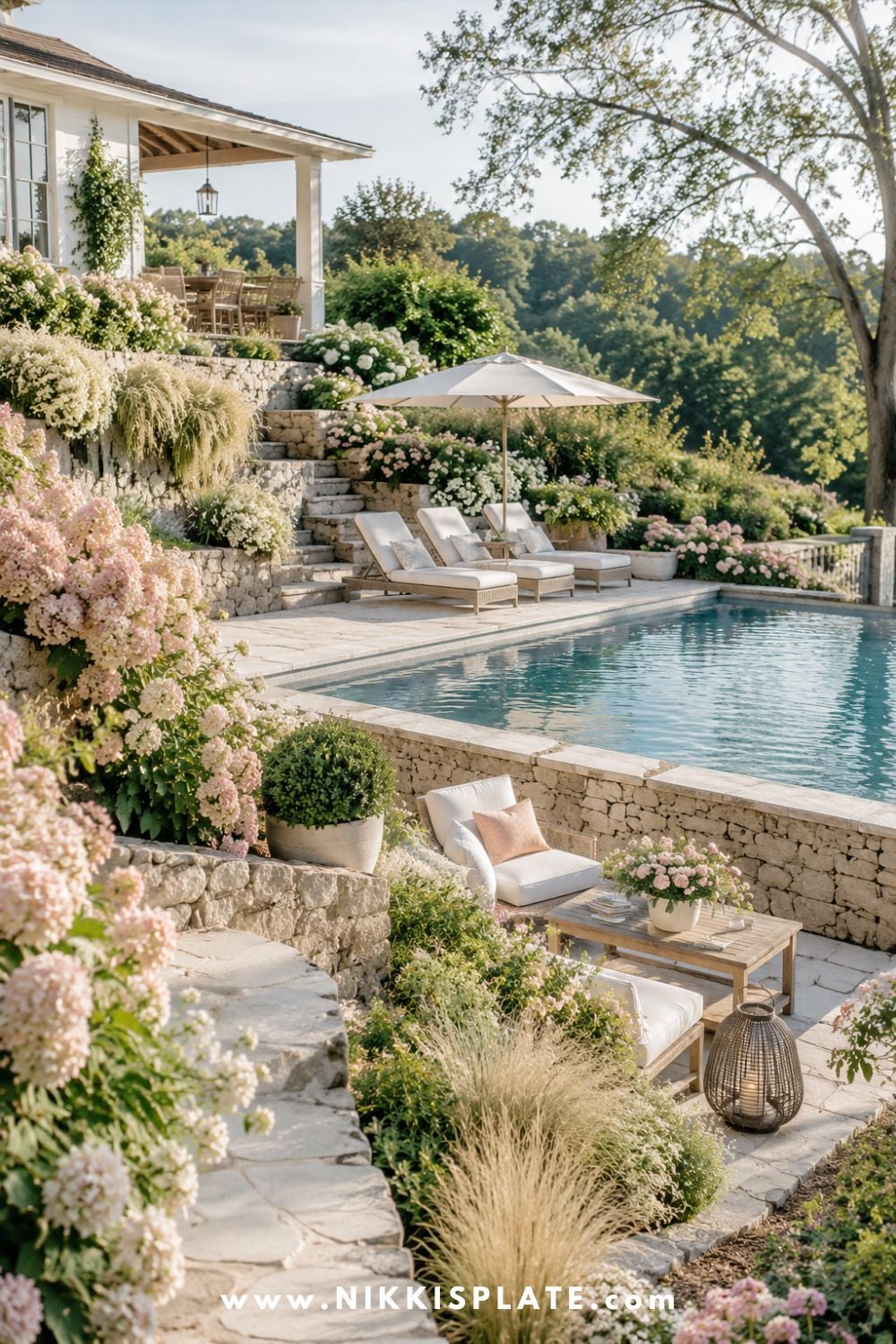 Sloped backyard landscaping with a raised pool area, stone retaining walls, lounge chairs, soft cottage flowers, and layered hillside greenery.