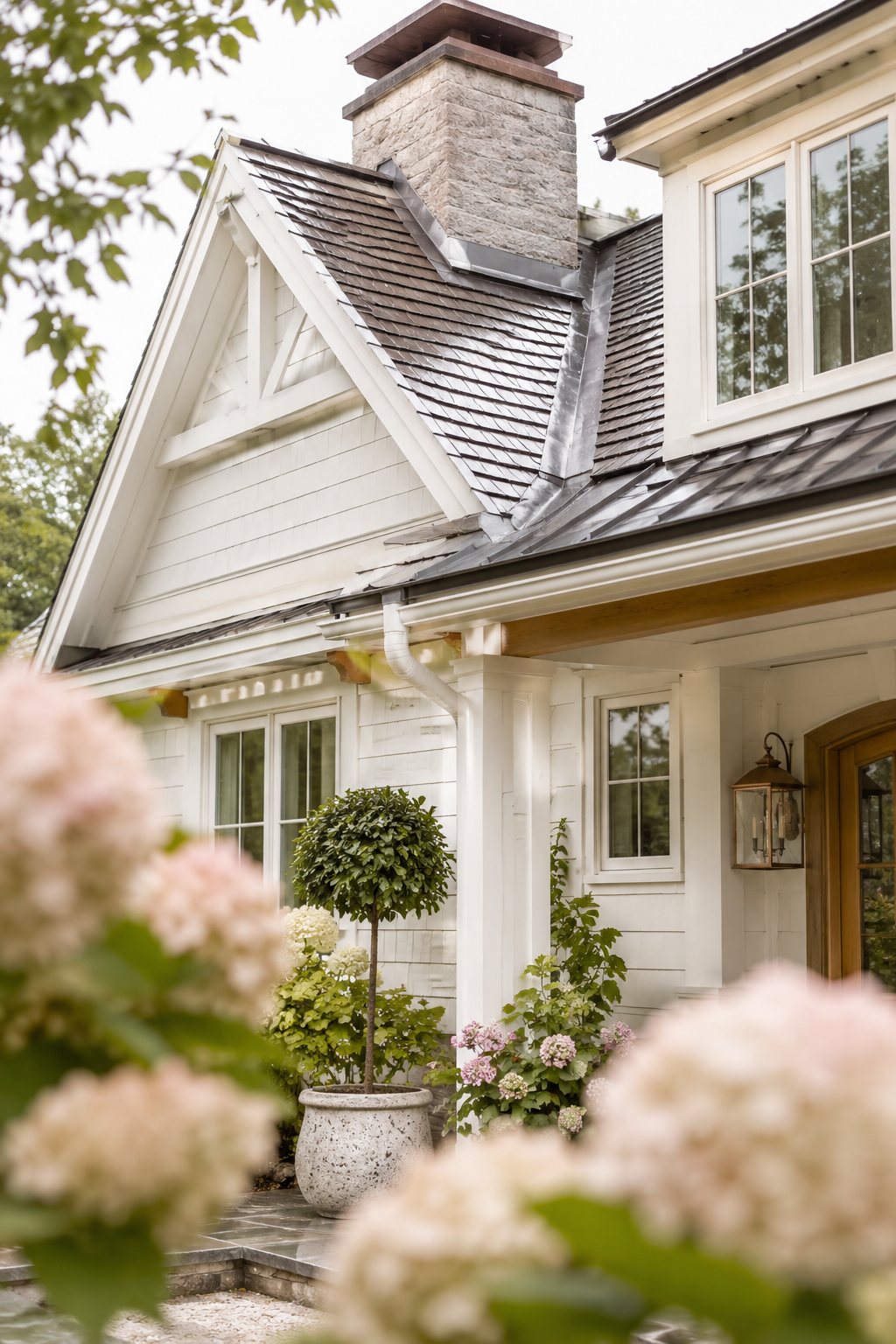 Soft farmhouse exterior with a clean roofline, chimney flashing, gutters, and neutral shingles framed by hydrangeas after a light rain.