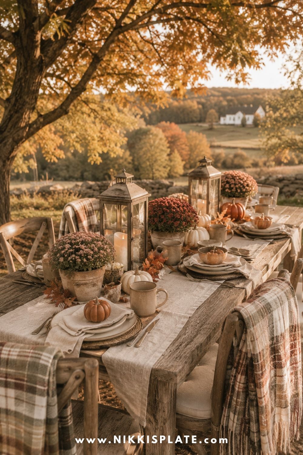 Outdoor fall table setting with pumpkins, lanterns, potted mums, cream linens and plaid throws beneath autumn trees.