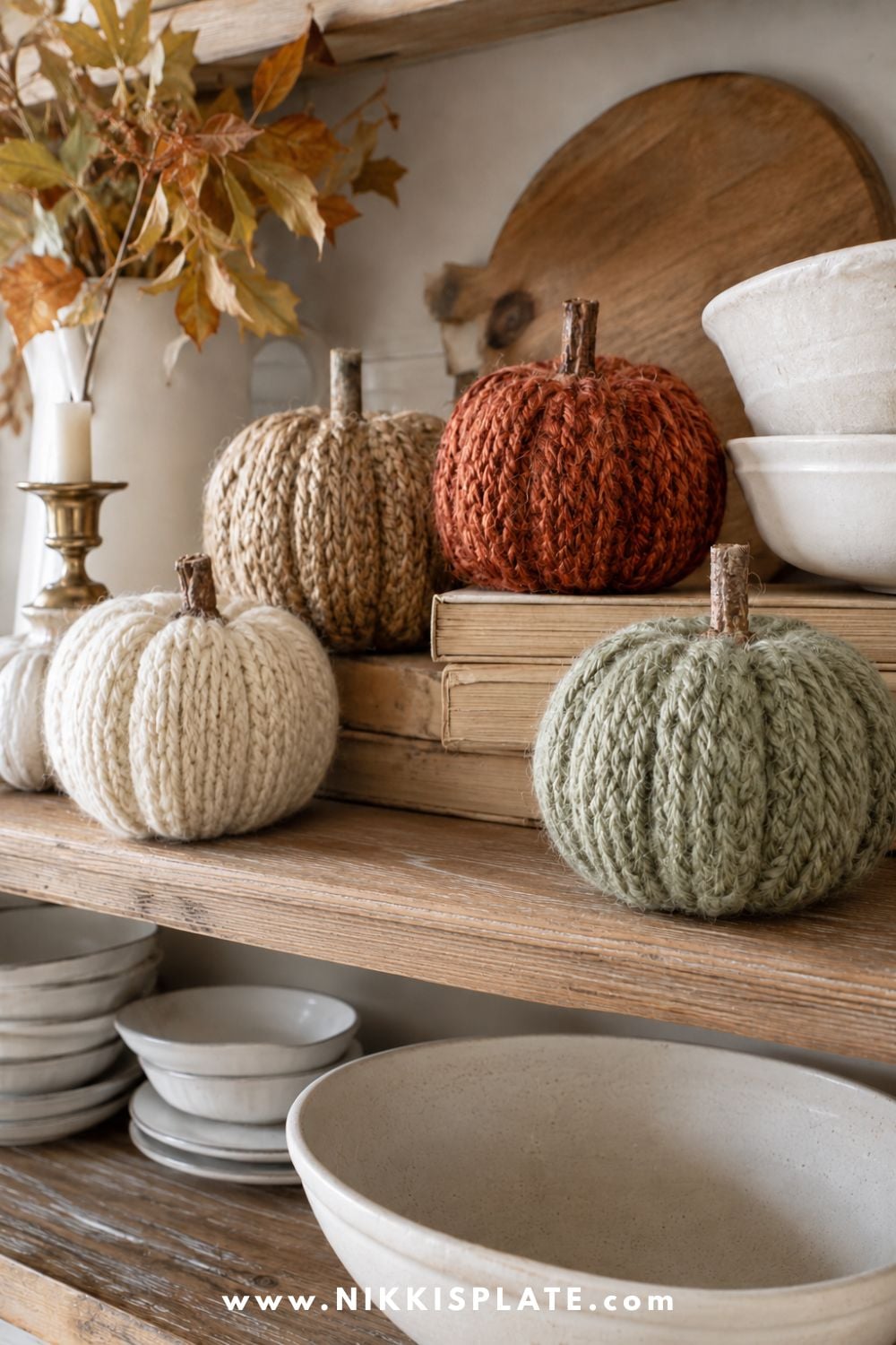 Chunky yarn-wrapped pumpkins in cream, caramel, rust and sage displayed with vintage books on open farmhouse shelves.