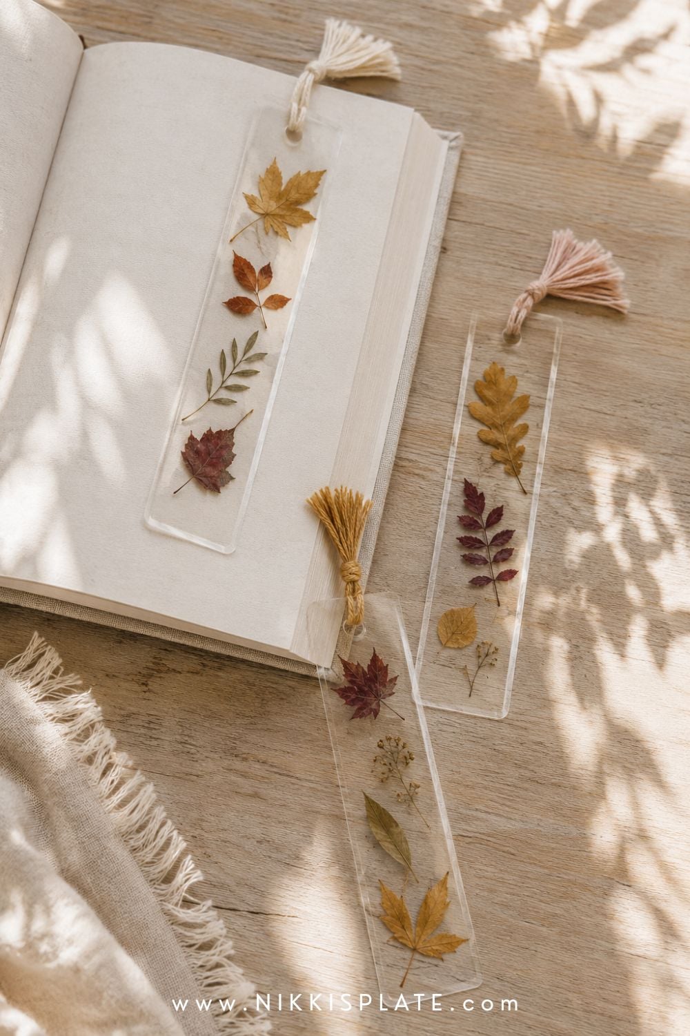 Pressed leaf bookmarks with tiny autumn leaves and cream, blush and mustard yarn tassels beside a book.