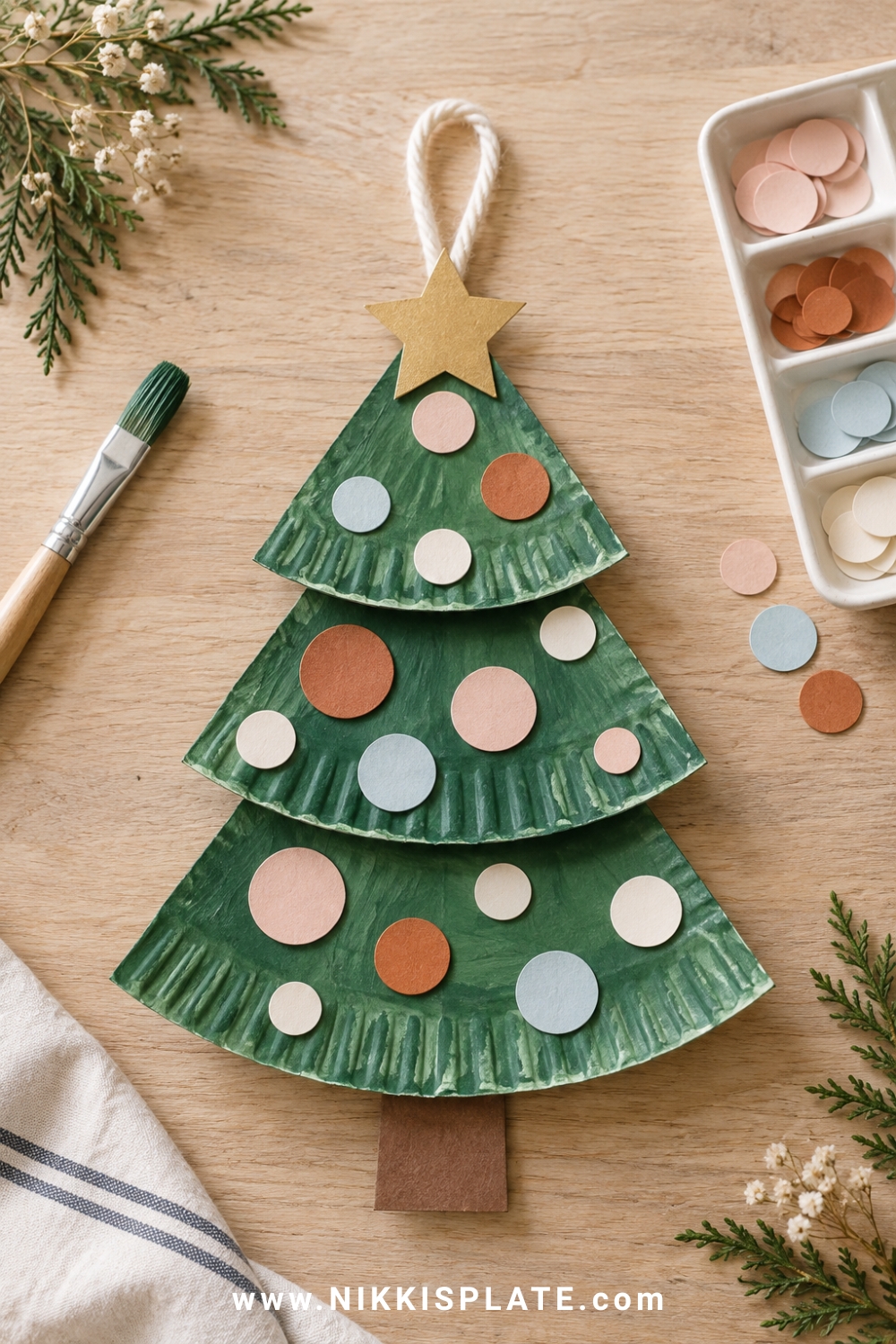 Green paper plate Christmas tree with pastel paper ornaments, gold star and yarn loop on a pale oak table.