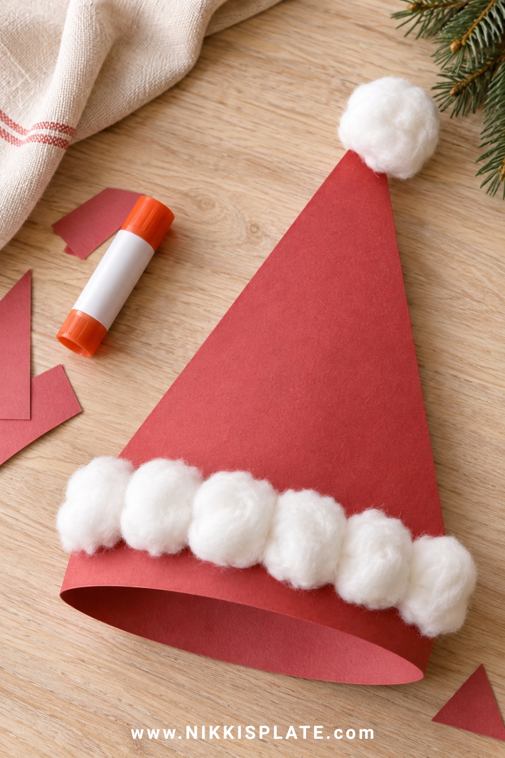 Red cardstock Santa hat craft trimmed with fluffy white cotton balls on a pale oak table.