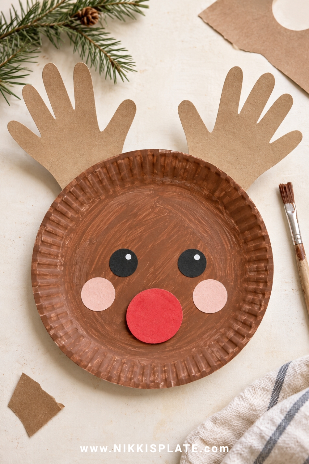 Brown paper plate reindeer with handprint antlers, rosy cheeks and a red paper nose.