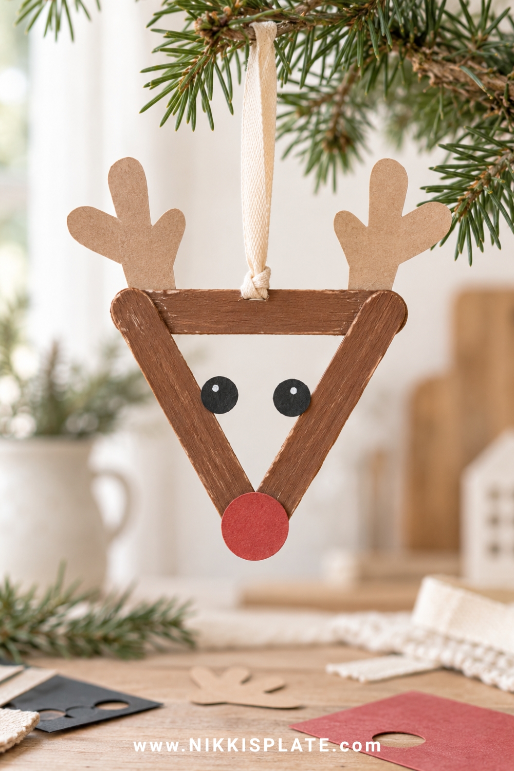 Popsicle stick reindeer ornament with kraft paper antlers, red nose and cream ribbon.