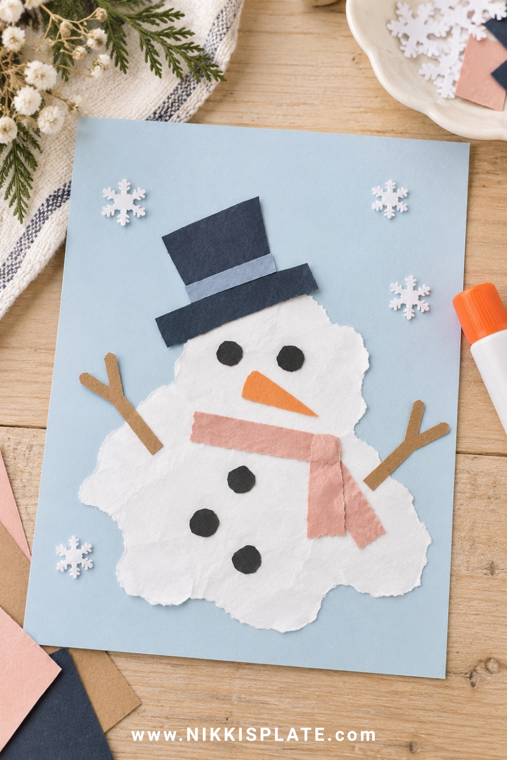 Melted snowman paper collage with crooked hat, carrot nose and scattered buttons on blue cardstock.