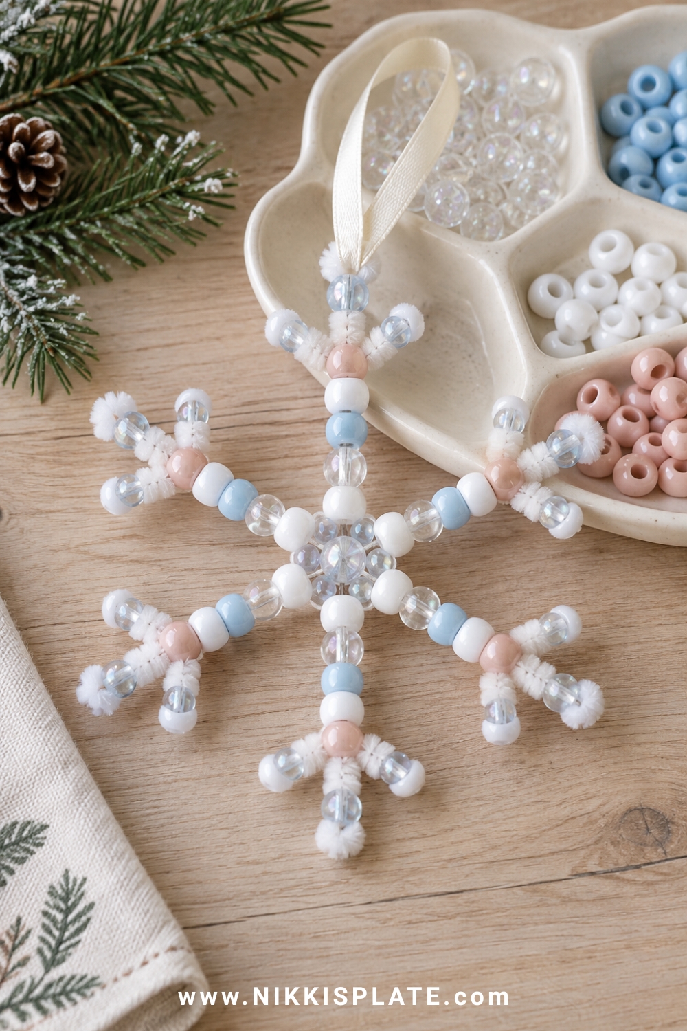 A beaded pipe cleaner snowflake ornament with clear, white, pale blue and blush pony beads arranged on a small ceramic sorting tray, cream ribbon hanger.