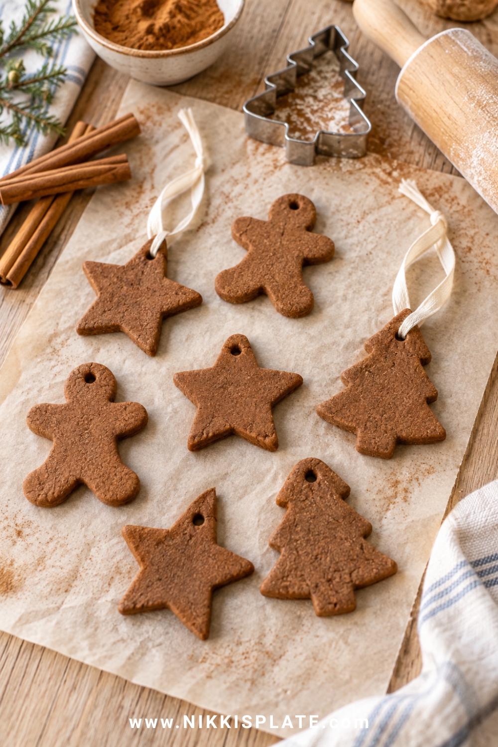 Cinnamon applesauce ornaments shaped like stars, trees and gingerbread people with cream ribbon, drying on parchment beside cinnamon sticks and a small rolling pin.