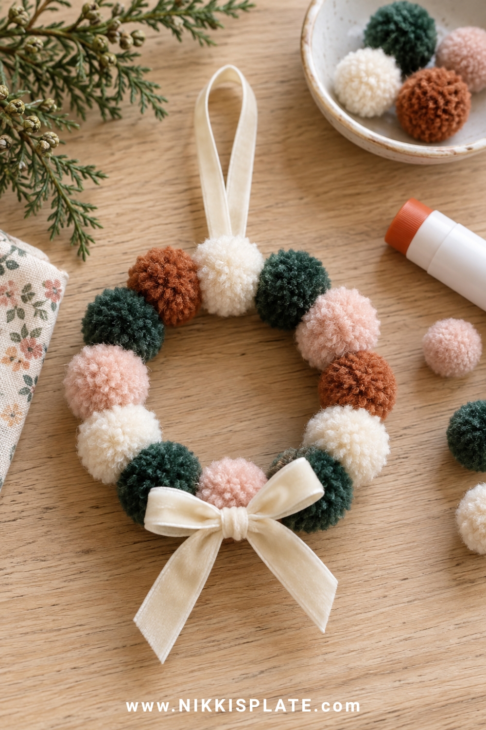 A small pom pom wreath ornament in muted green, cream, blush and rust with a velvet bow and ribbon loop, drying flat on a pale oak craft table.