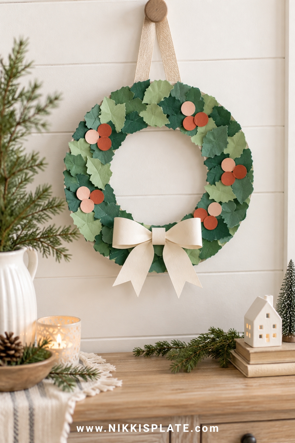 A paper plate christmas wreath covered in layered torn green paper with blush and rust paper berries, a cream bow and natural ribbon hanger.