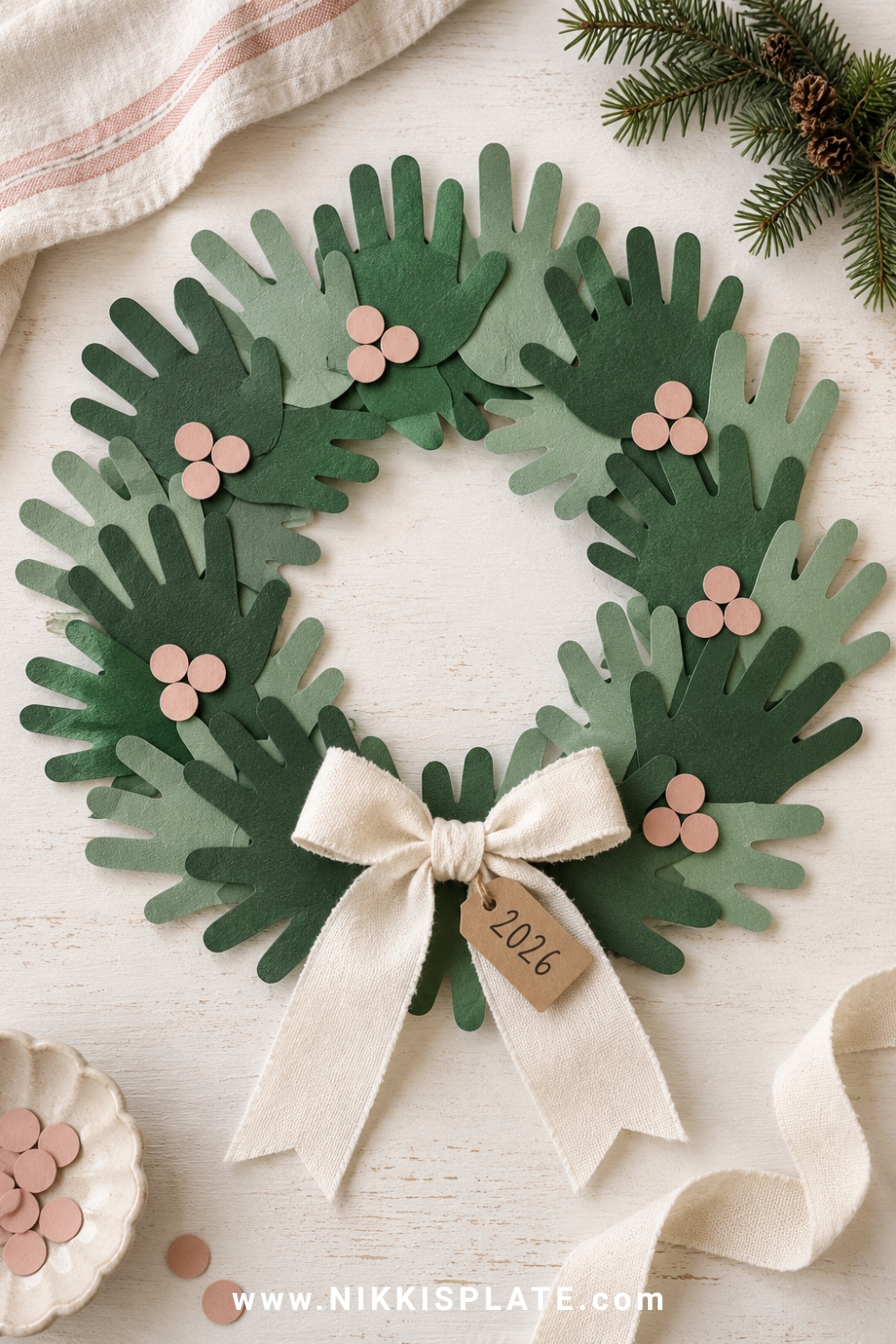 A layered handprint christmas wreath made from two shades of green cardstock with blush paper berries, a soft cream bow and a handwritten family year tag.