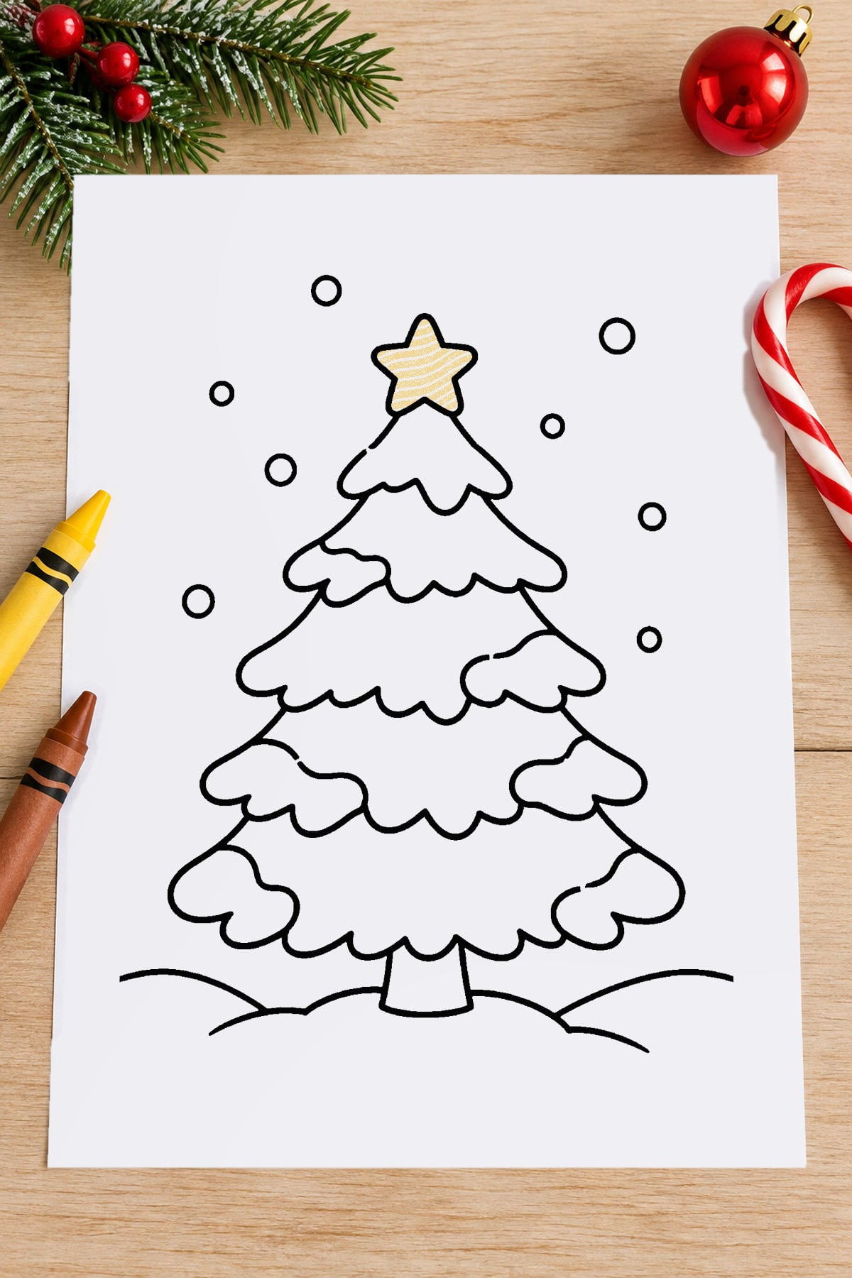 Snow-covered Christmas tree coloring page with a yellow star and falling snowflakes, displayed on a Christmas-themed wooden table.