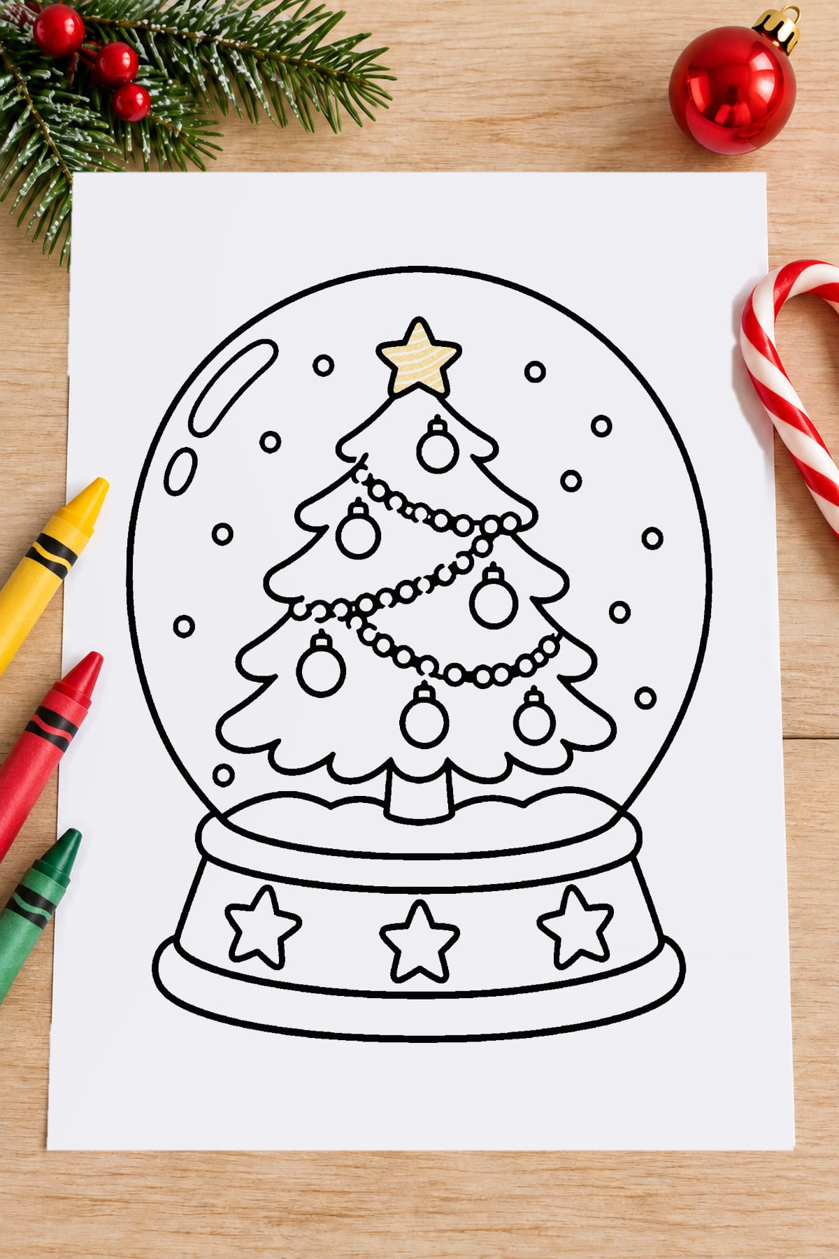Printable Christmas tree snow globe coloring page featuring a decorated tree, falling snow, and star-covered base on a wooden craft table.