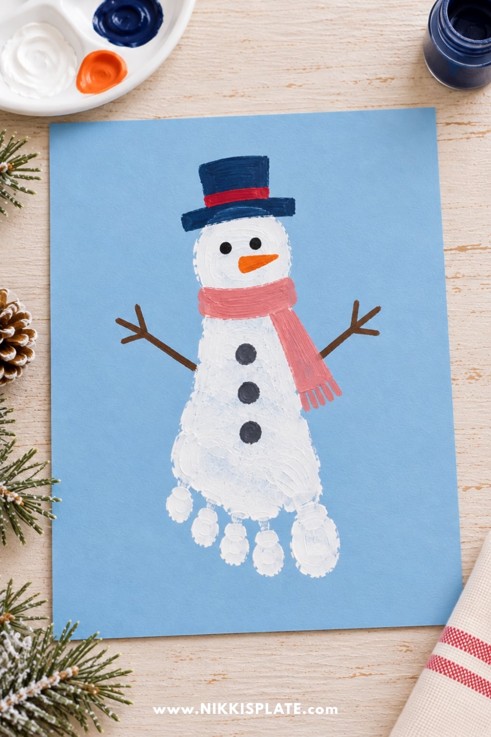 Cute footprint snowman craft for kids made with white paint on blue paper, featuring a navy hat, pink scarf, and carrot nose.