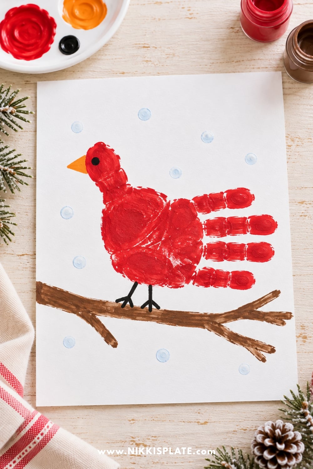 Red handprint Christmas cardinal craft for kids with an orange beak, black eye, and painted branch surrounded by falling snow.