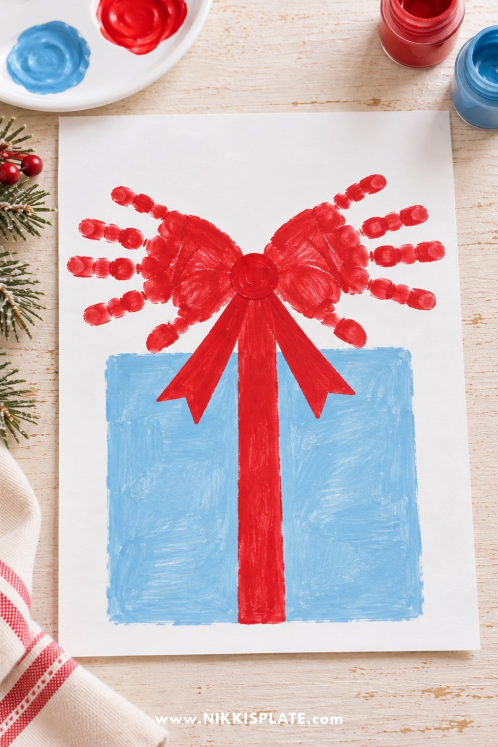 Easy Christmas handprint craft featuring two red handprints forming a festive bow on a blue Christmas present with red ribbon.