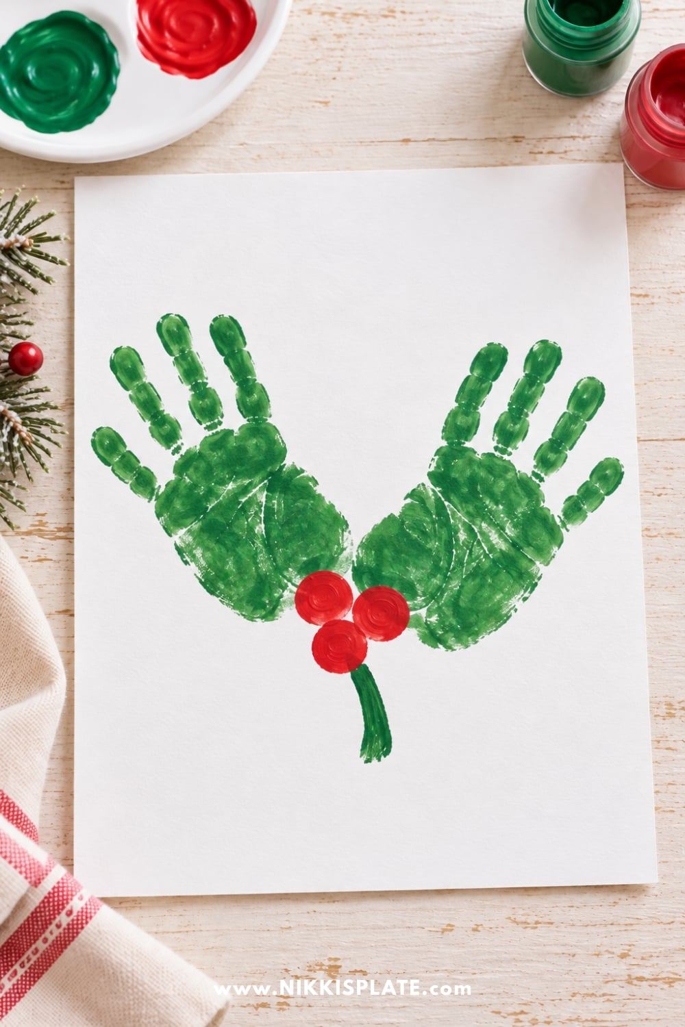 Easy Christmas handprint holly craft for kids featuring two green painted handprints, three red fingerprint berries, and a green stem.