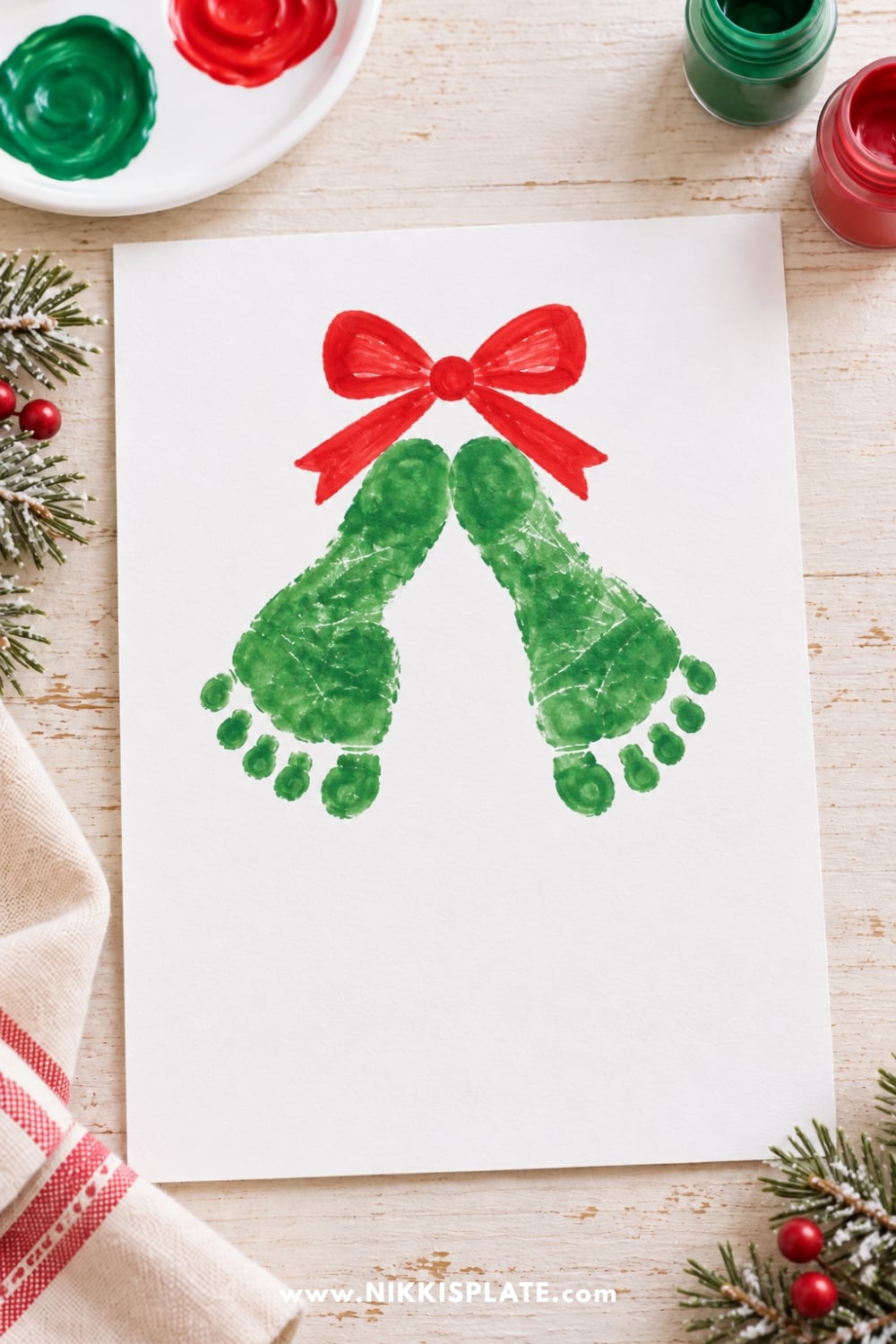 Christmas footprint keepsake for kids featuring two green painted footprints arranged like mistletoe beneath a festive red bow.