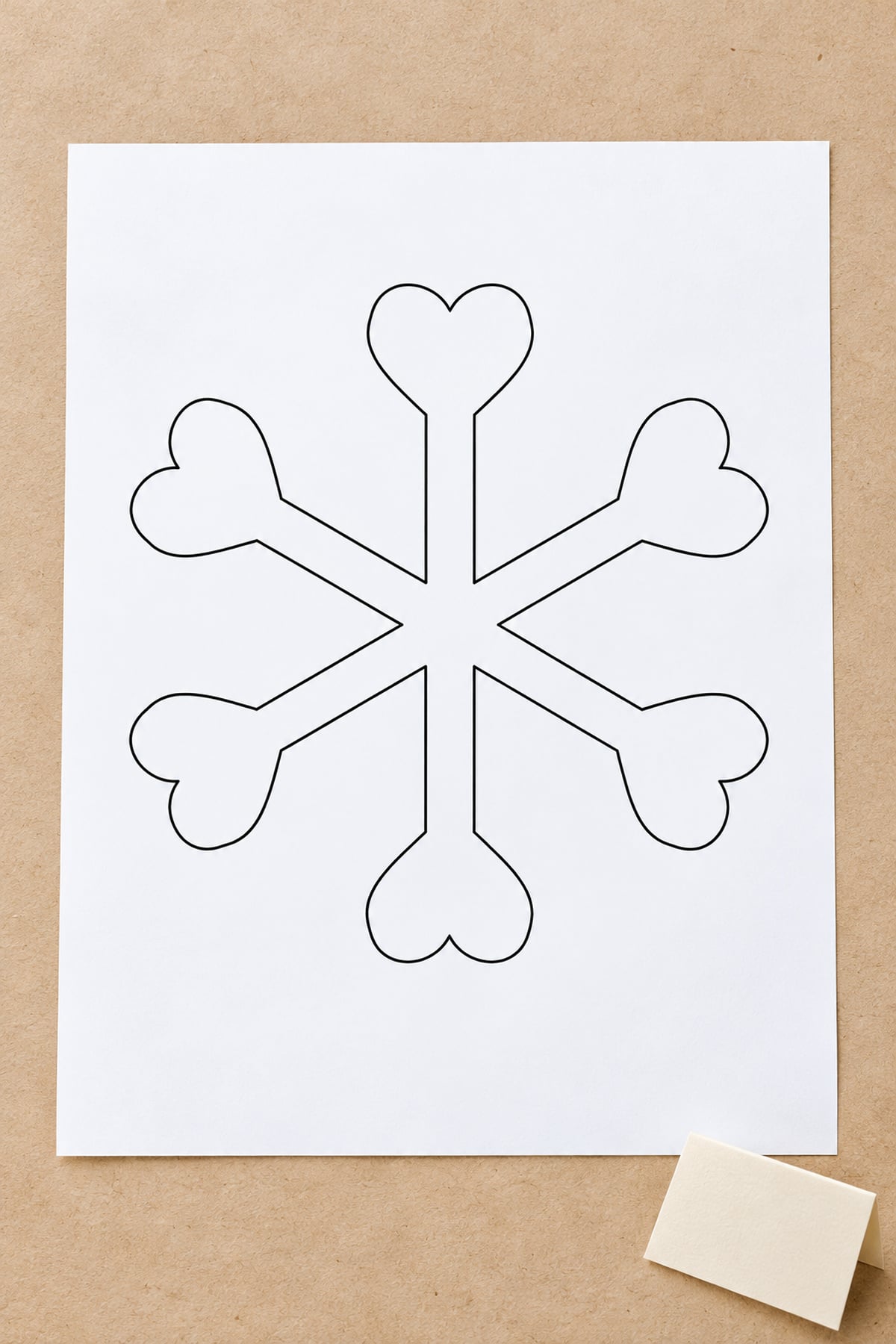 Heart-tip snowflake outline on white paper over kraft paper beside a blank cream card.