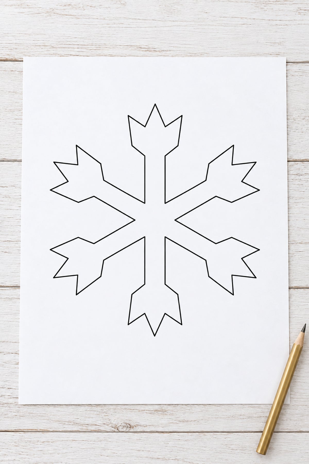 Crown-tip snowflake outline on white paper on a whitewashed wooden table beside a gold pencil.
