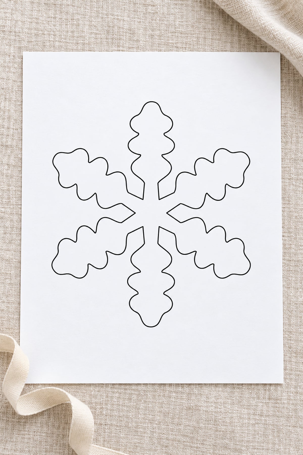 Oak-leaf snowflake template on white paper over oatmeal linen, with a cream cotton ribbon.