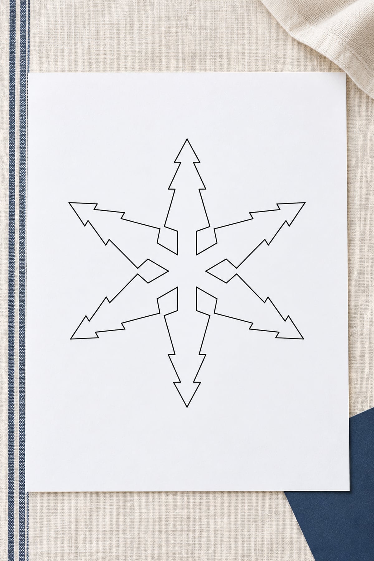 Evergreen-branch snowflake template on an ivory cloth with navy stripes and a navy paper corner.