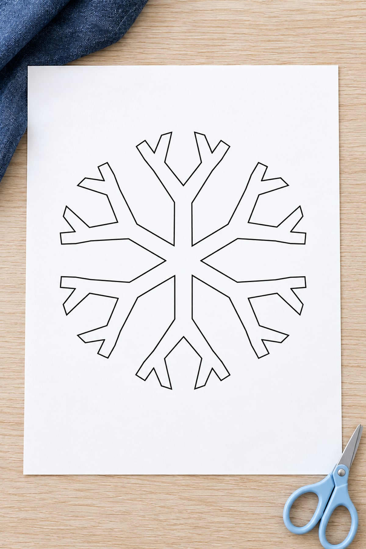 Double-fork snowflake template on a pale wood table with navy fabric and blue scissors.