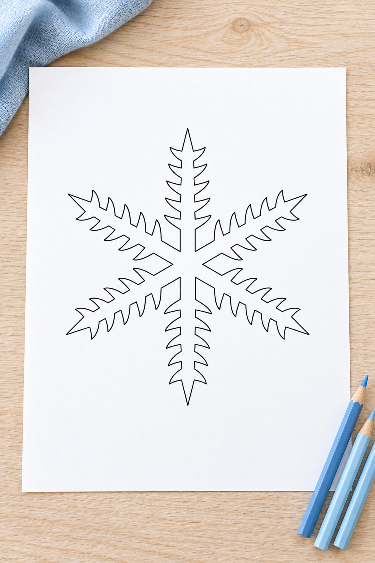 Fern-frond snowflake outline on a light wood table with pale blue fabric and blue pencils.