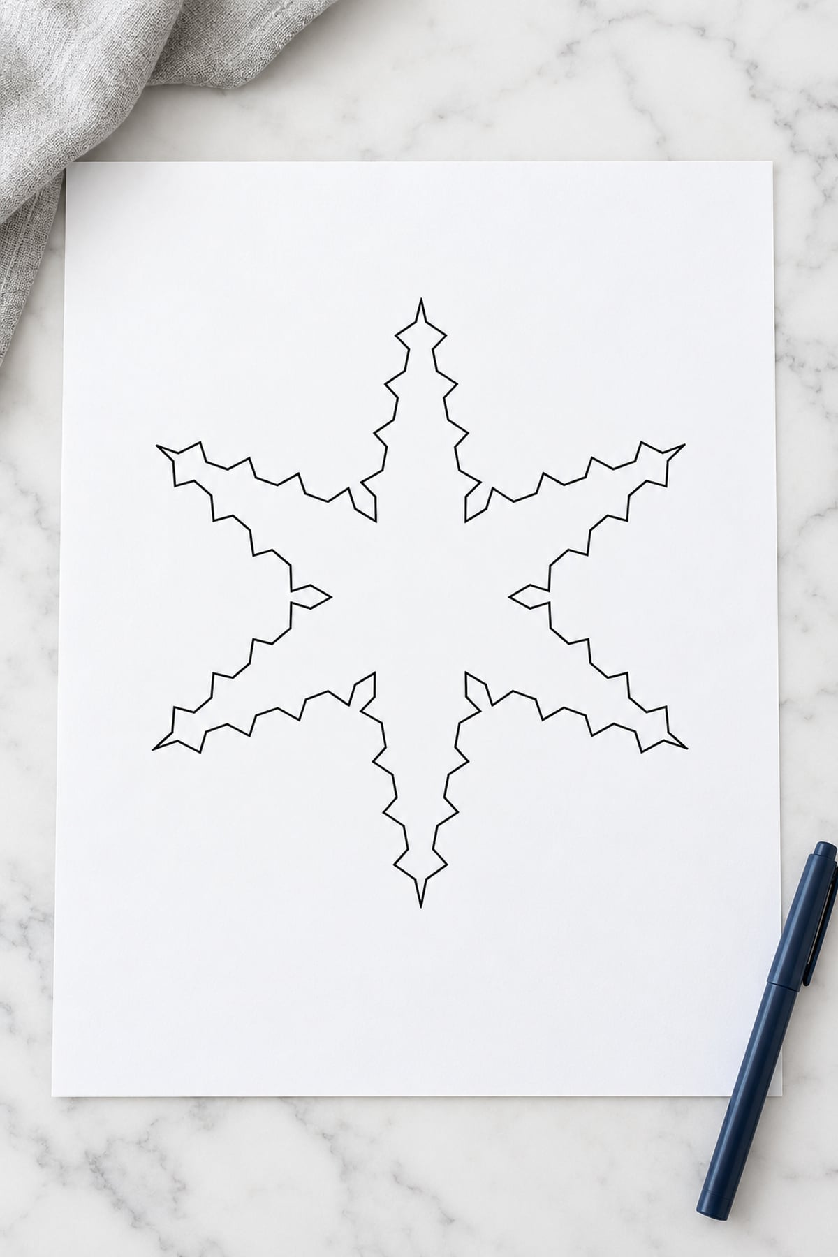 Sawtooth-edge snowflake outline on white marble with grey linen and a navy pen.