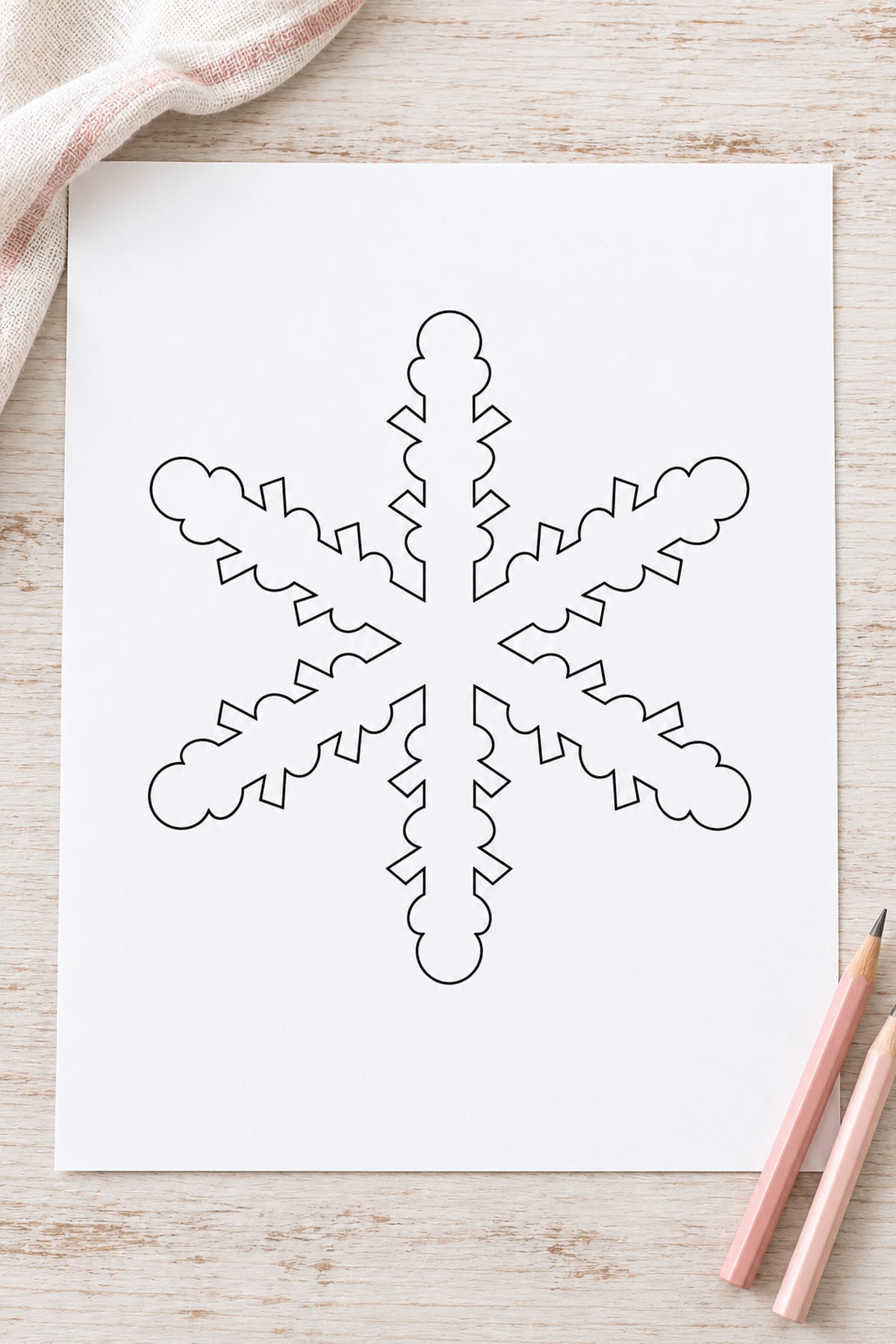 Lace-edge snowflake outline on whitewashed wood with blush-striped ivory linen and pink pencils.