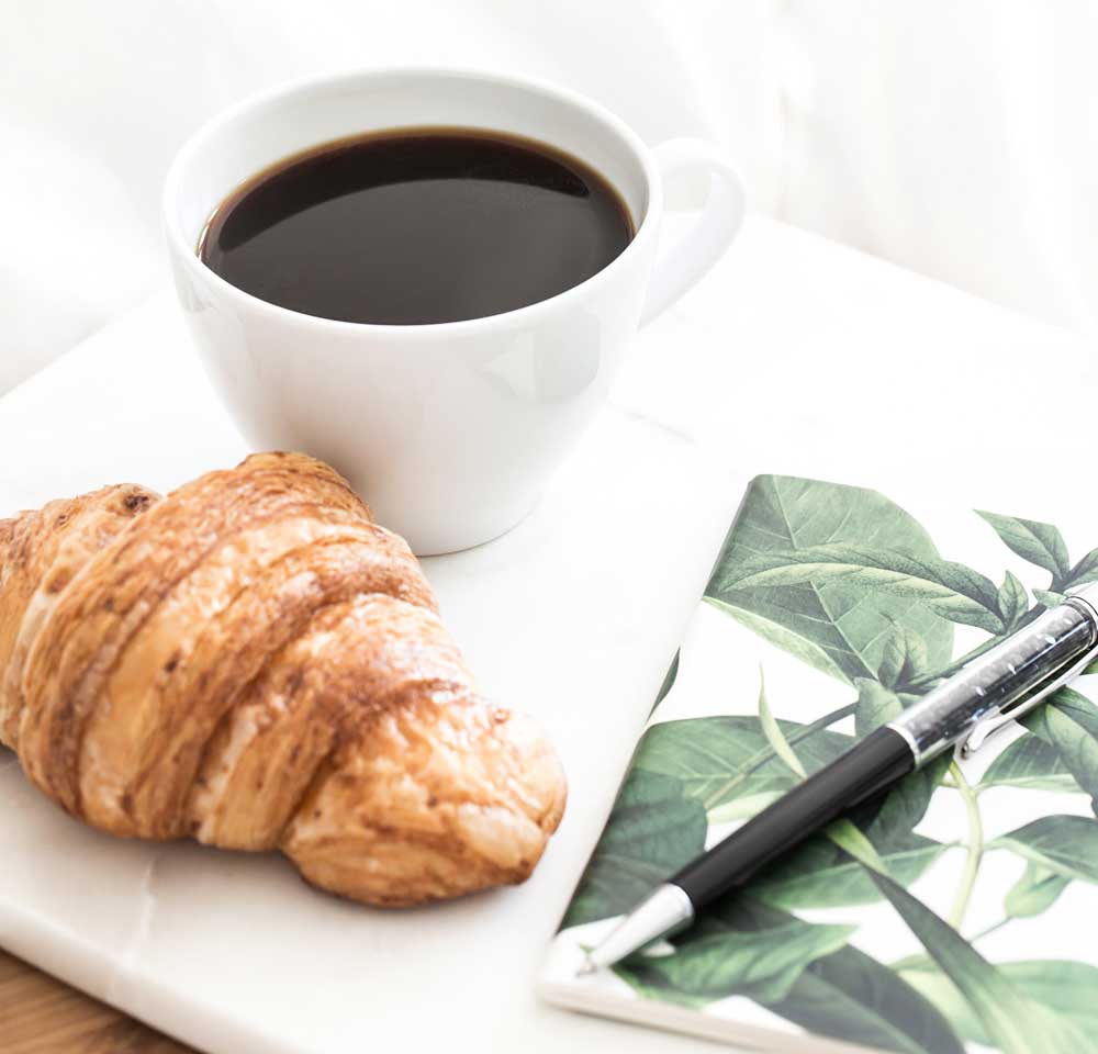 A cup of black coffee, a croissant, and a notebook and pen on a white plate