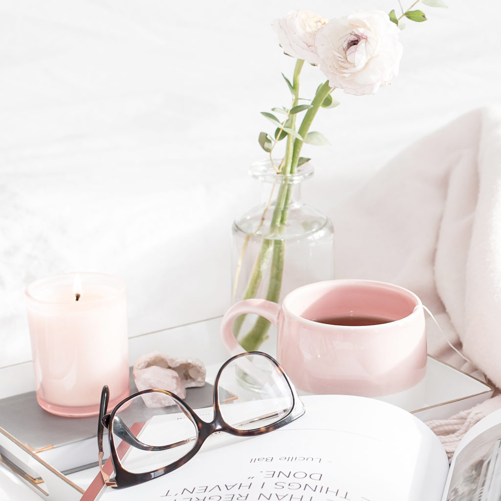 Bring more positivity into your life with these tip! (image: flowers in a vase, a pink mug with tea, a candle, reading glasses and a book on a table)