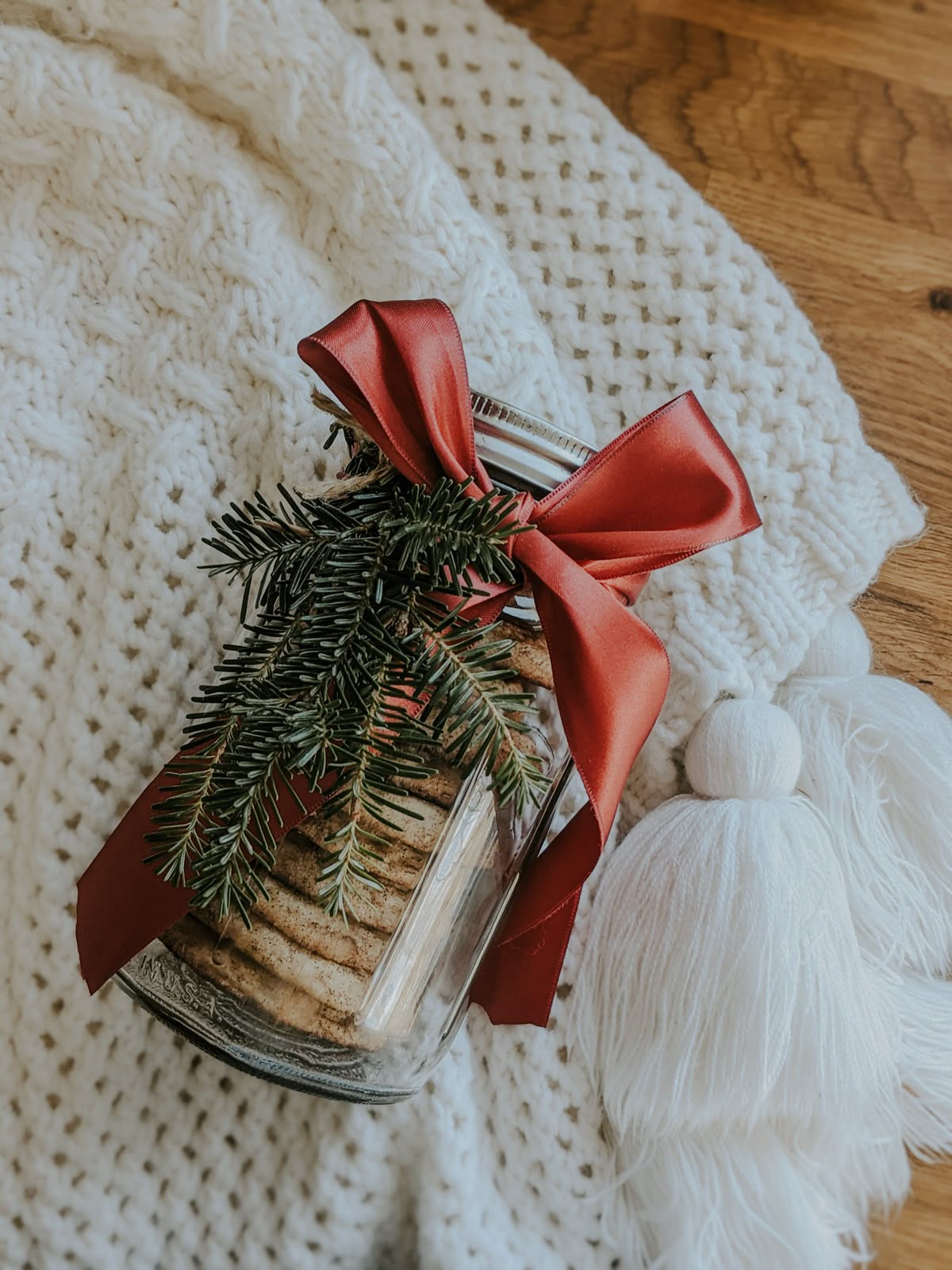 Homemade Christmas cookies stacked in a Mason jar with a red ribbon and evergreen sprig, a festive DIY holiday gift idea.