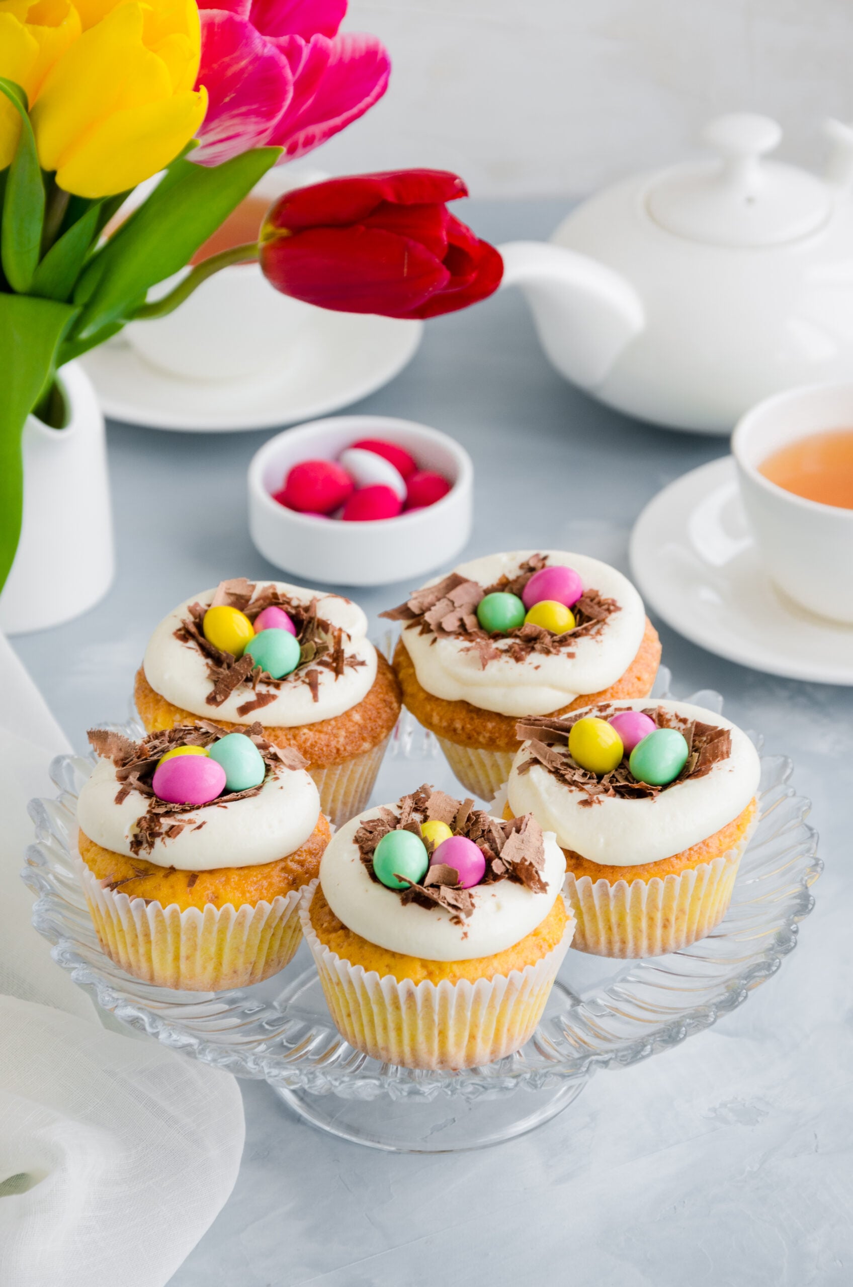 Bird's nest vanilla cupcakes with white frosting and pastel candies on a glass cake stand with tulips in the background