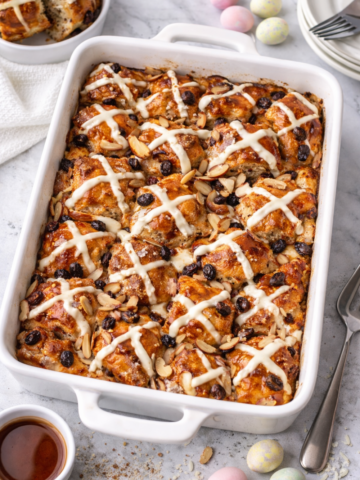 Hot cross bun French toast casserole baked in a white dish with icing drizzle, raisins, and sliced almonds