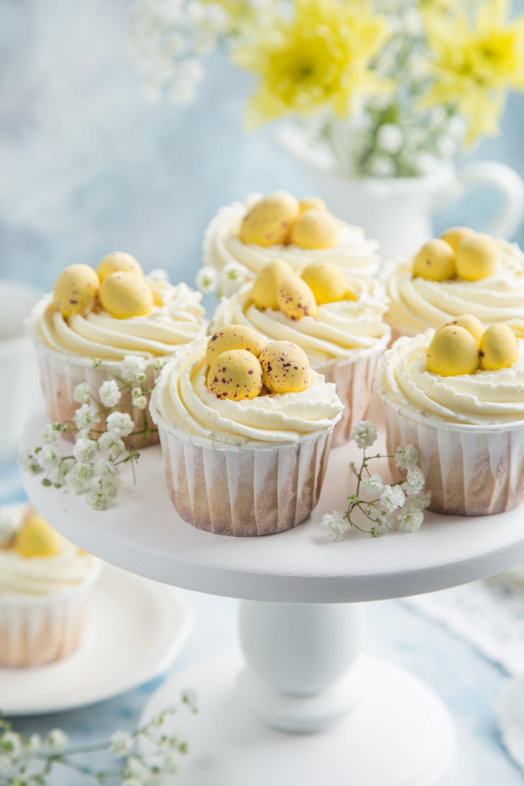 Vanilla Easter cupcakes with creamy frosting and yellow candy eggs, styled on a white cake stand with spring flowers